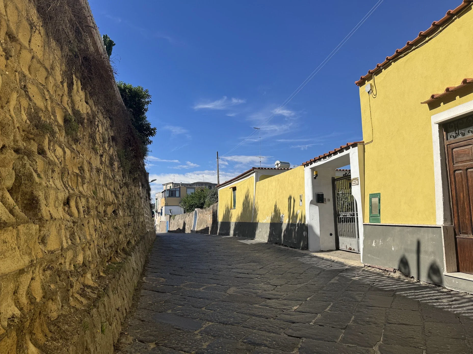 A narrow cobblestone street with a stone wall on the left and yellow houses on the right, under a clear blue sky.