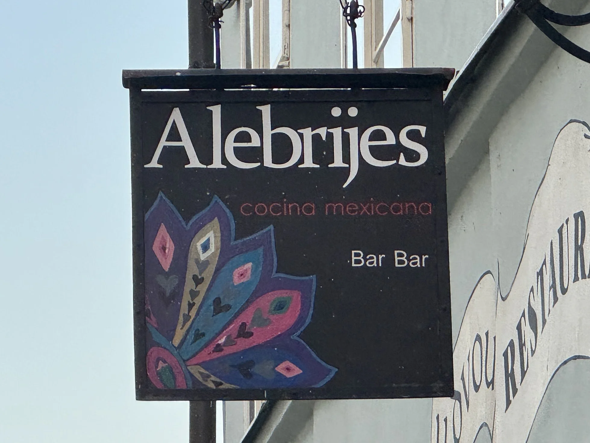 Sign for a Mexican restaurant called Alebrijes with colorful artwork on it, located outdoors.