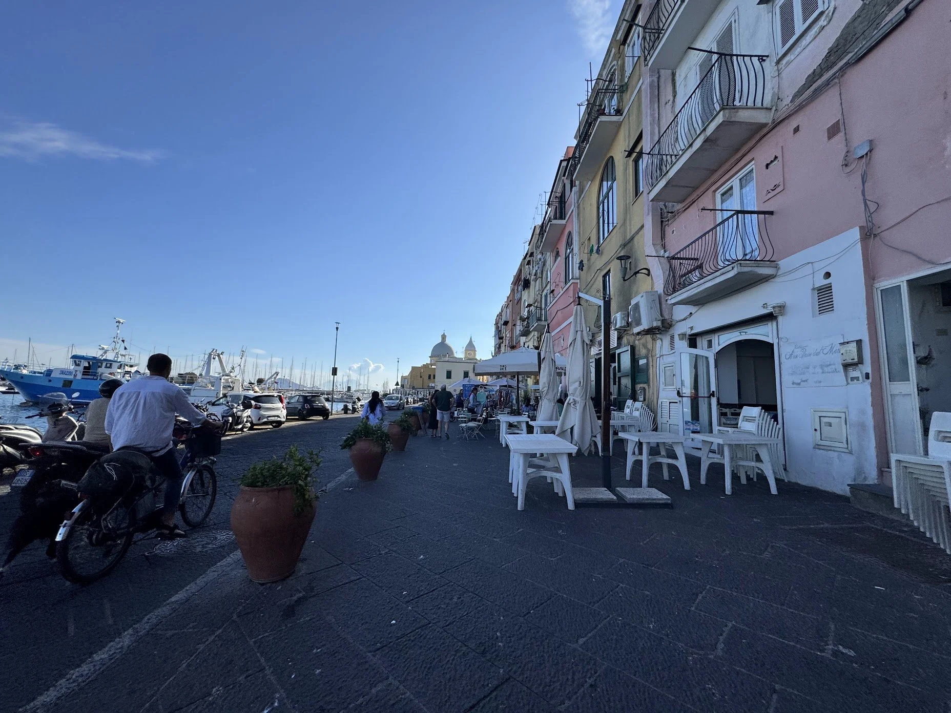 Street view with colorful buildings, outdoor seating, people walking, and boats docked at the marina in the background under a blue sky.