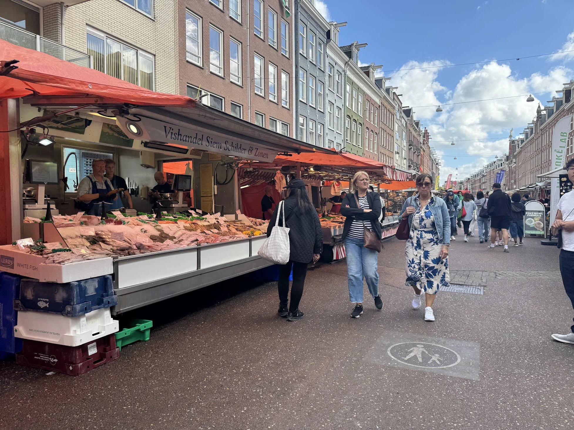 People walking along a street market with various stalls, some with seafood and fish, in a city with colorful buildings and a blue sky with clouds.