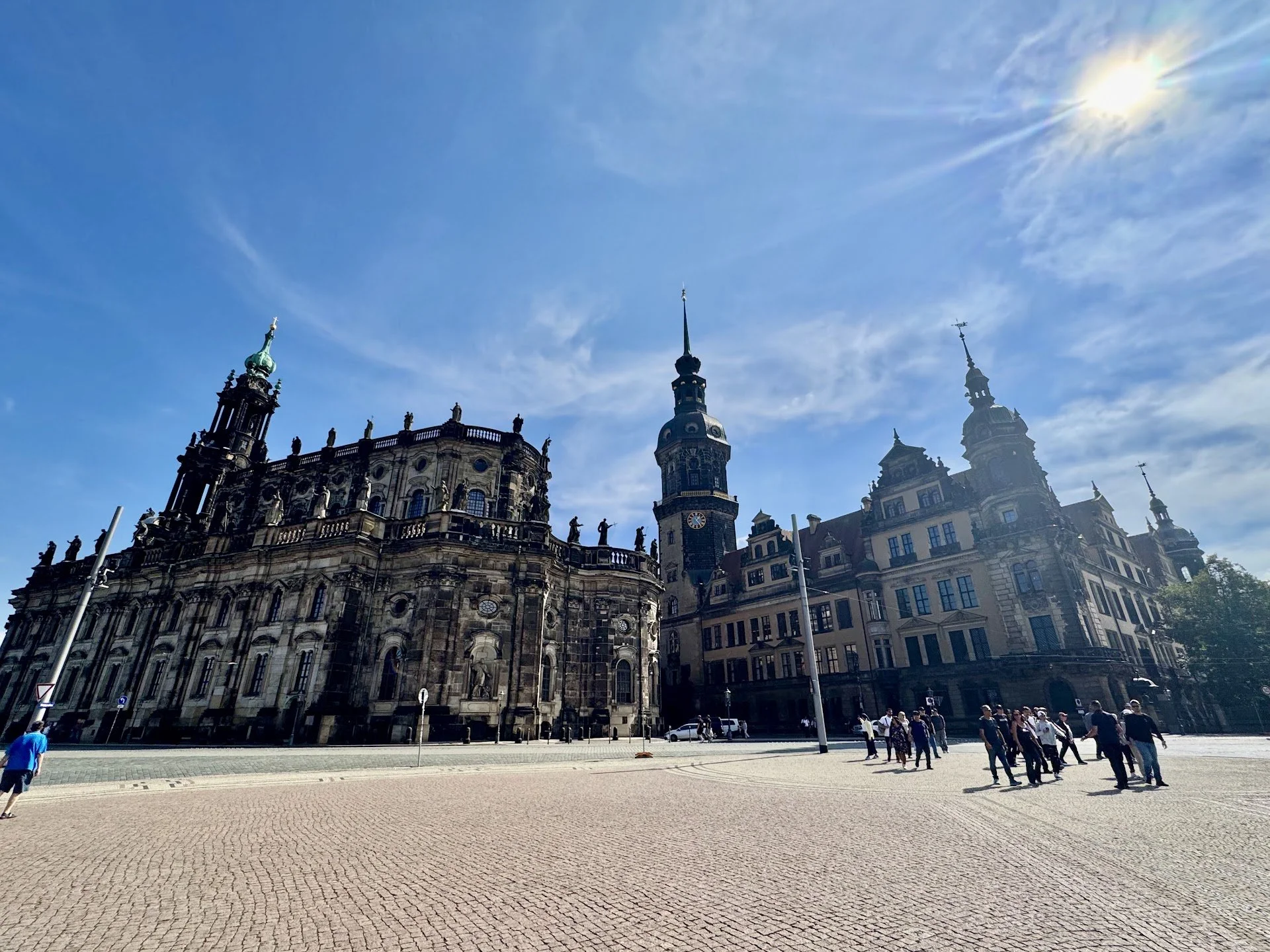 Historic European-style building with ornate architecture and clock tower, under a blue sky with the sun shining brightly, overlooking an open square with people walking.