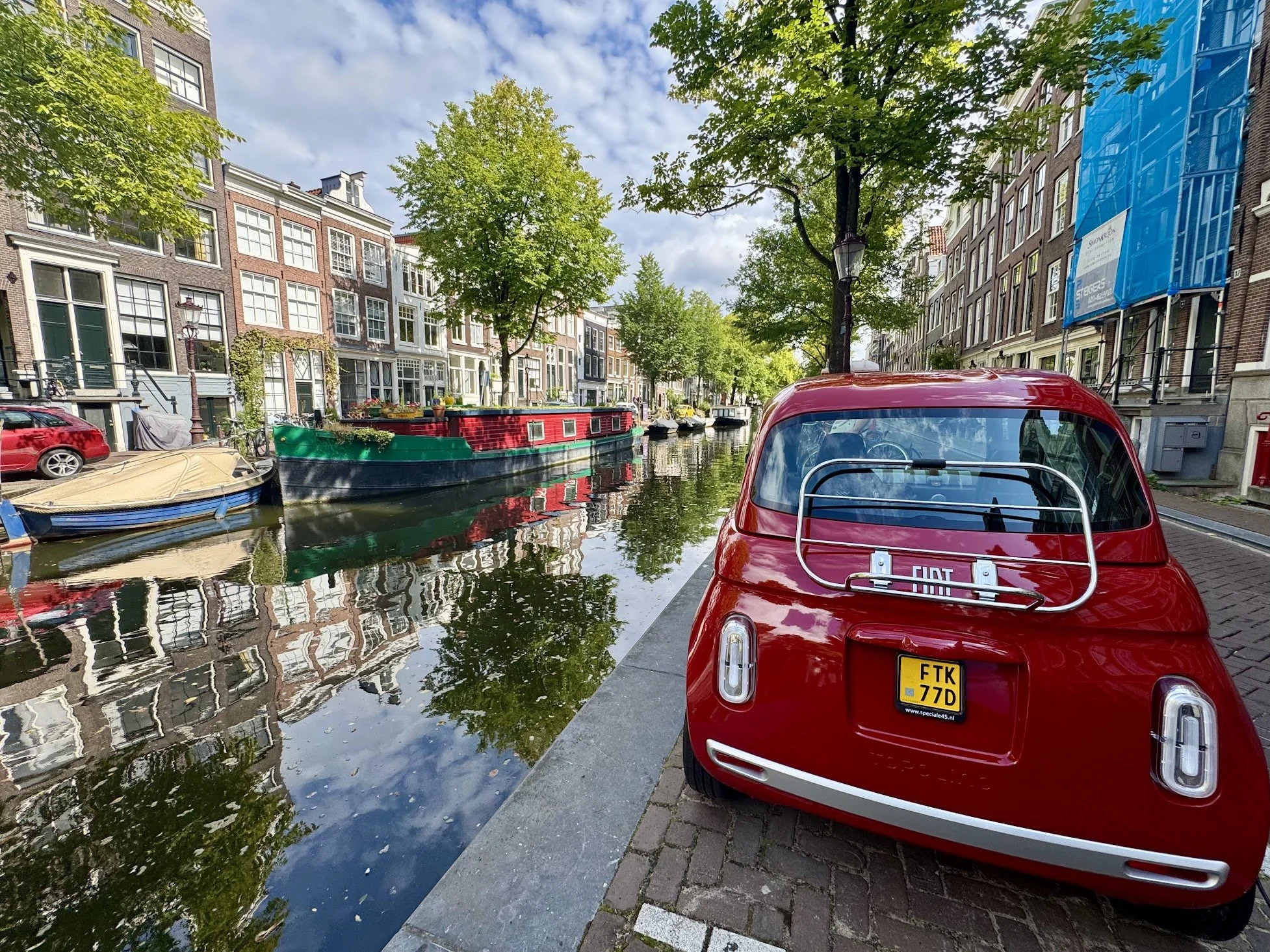 A canal with boat houseboats, trees, historic buildings, and cars parked along the street in Amsterdam. A red vintage Fiat car is in the foreground, and a reflection of the buildings can be seen in the canal water.