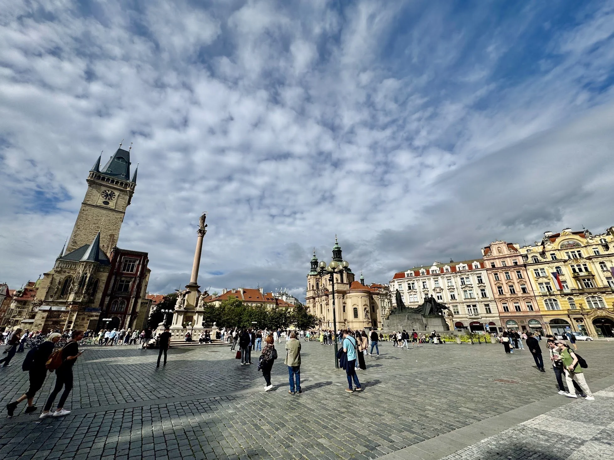 Public square in a European city with historic buildings, a tall clock tower, a column with a statue on top, and a crowd of people walking on cobblestone pavement.