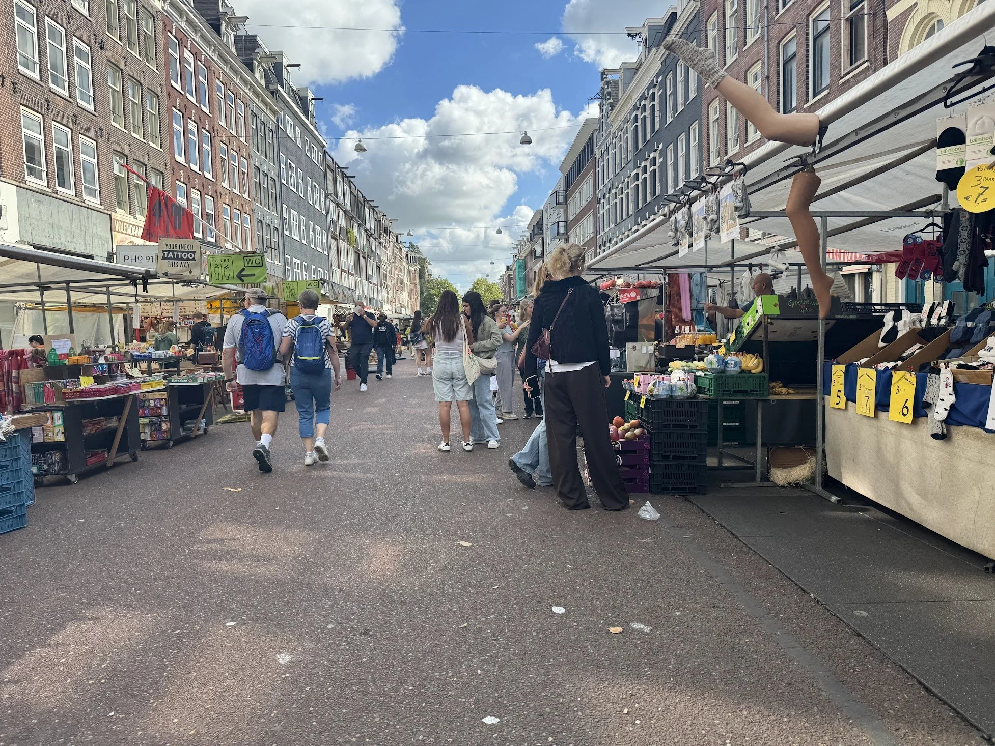 Street market with vendors selling goods, people browsing, brick buildings, tents, and a partly cloudy sky.