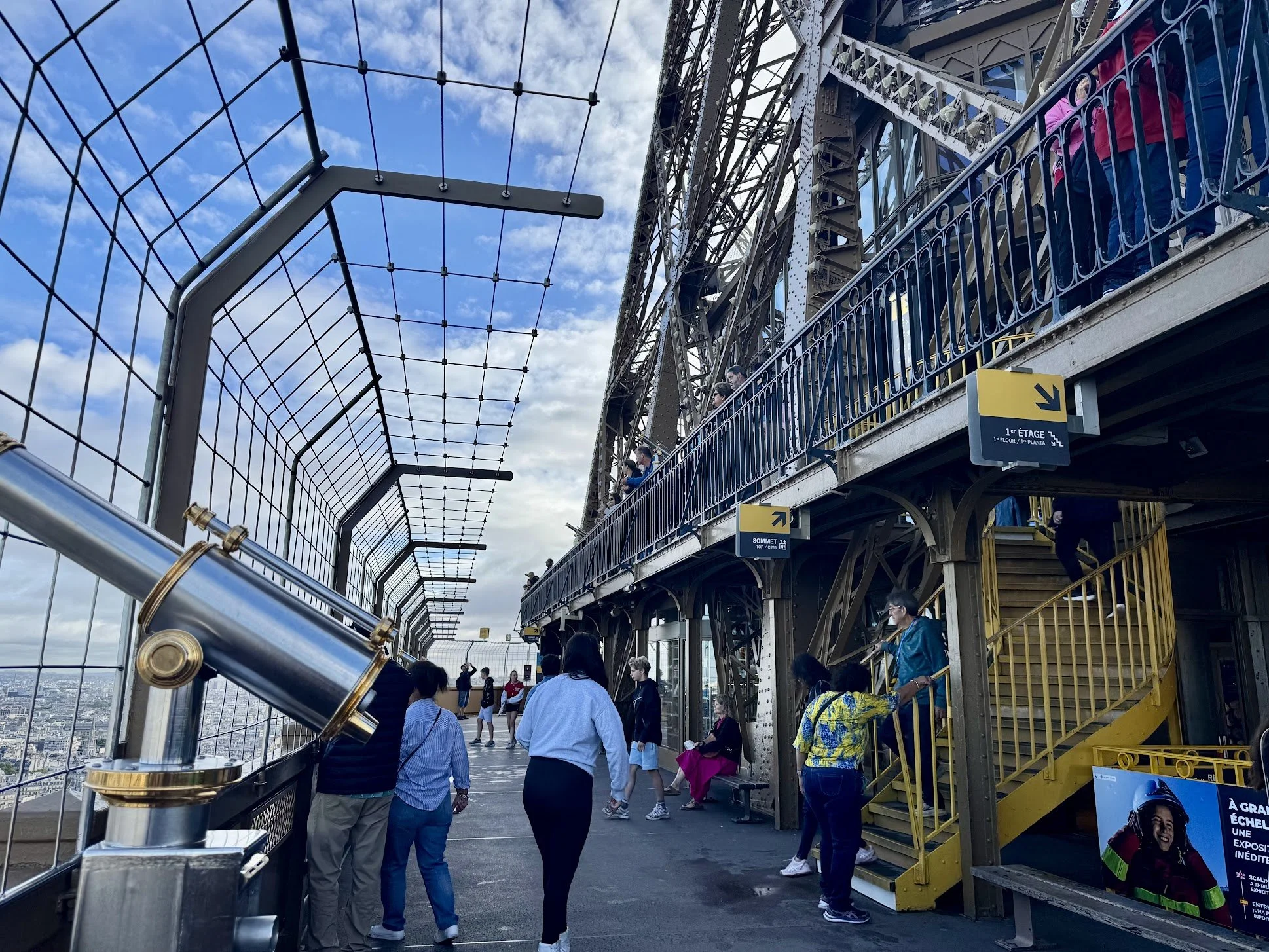 People standing on an observation deck or a similar high-rise structure with a view of a cityscape in the background. The deck is enclosed with safety fencing and metal railings, and there are signs in multiple languages indicating the different leve