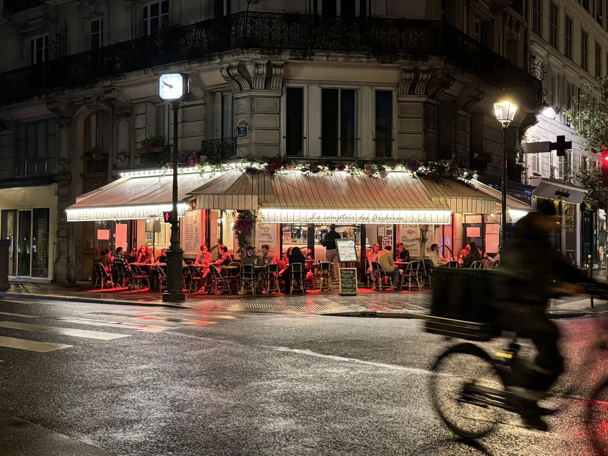 A Parisian street corner at night with a cafe called 'Le Comptoir des Archives,' outdoor seating filled with people, illuminated by street lamps and shop lights, wet street reflecting the lights, and a passing bicycle in motion.