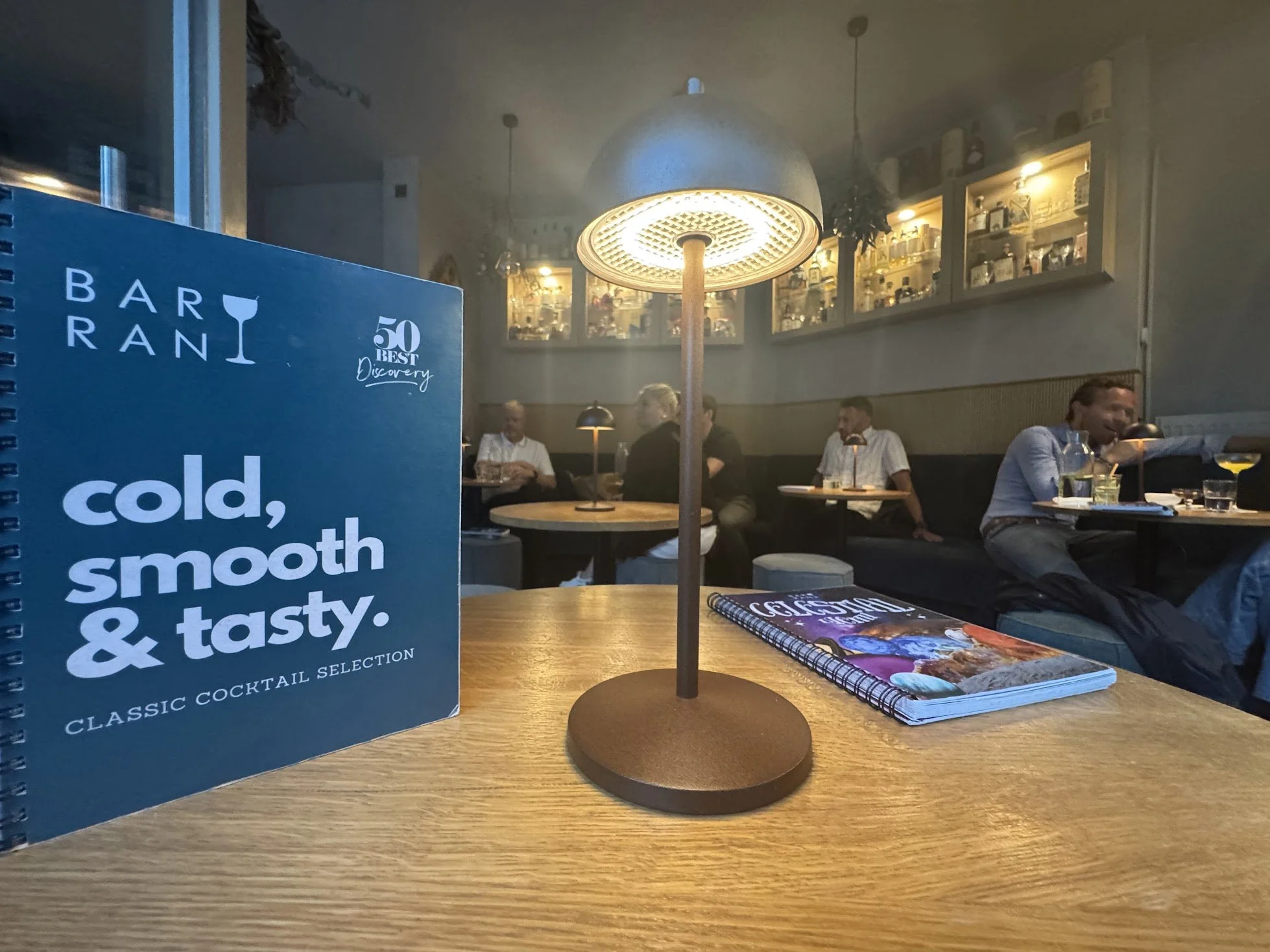 A restaurant interior with a blue menu on a wooden table, a decorative lamp, and several seated patrons in the background.