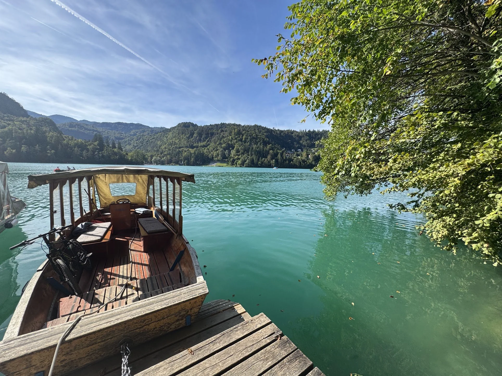 A wooden boat docked on a pier next to a lake with green water, surrounded by lush green trees and mountain hills in the background under a blue sky with streaks of clouds.