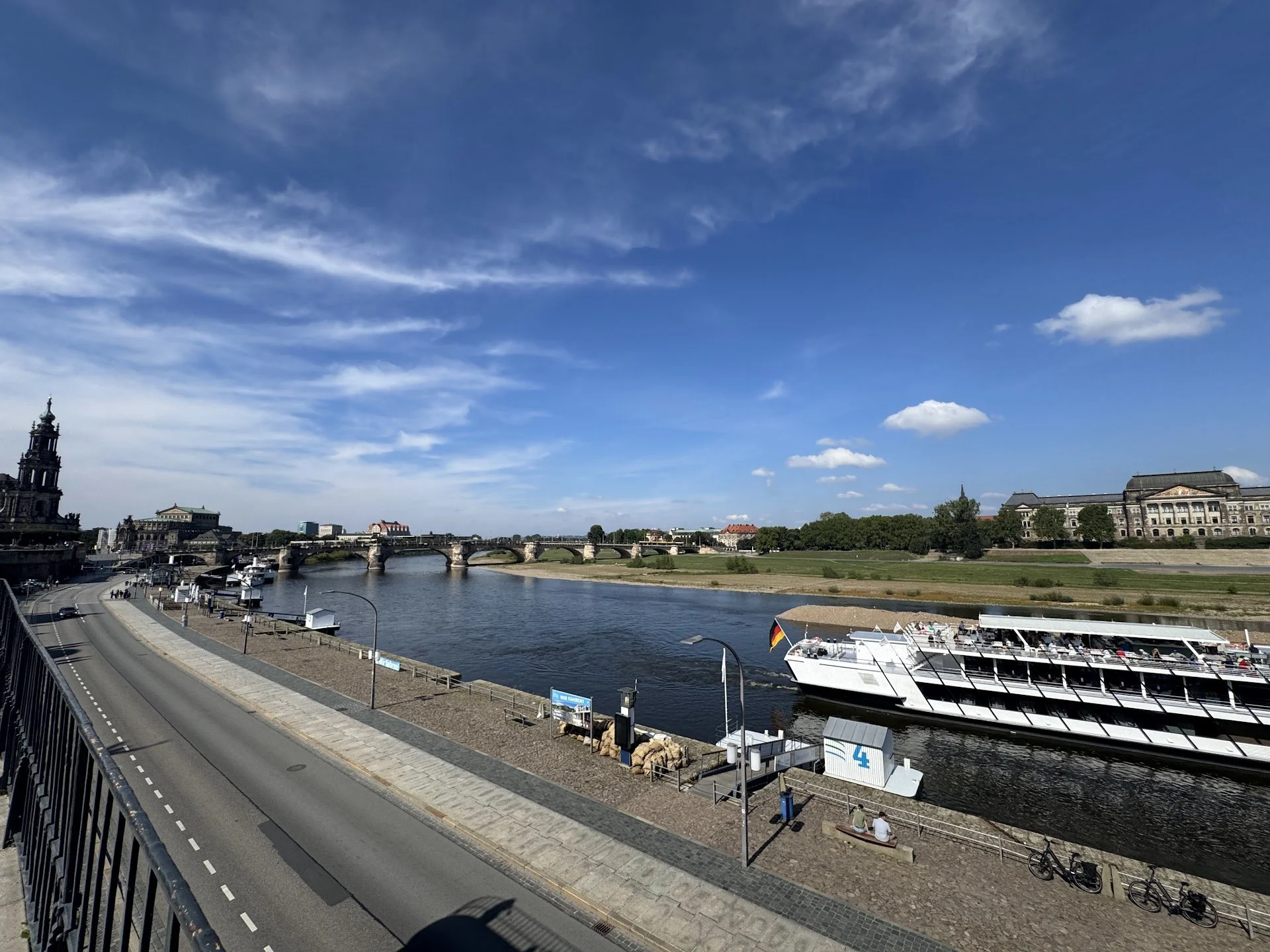 View of a river with a bridge crossing over it, city buildings in the background, and boats docked along the riverbank on a sunny day with blue sky and clouds.