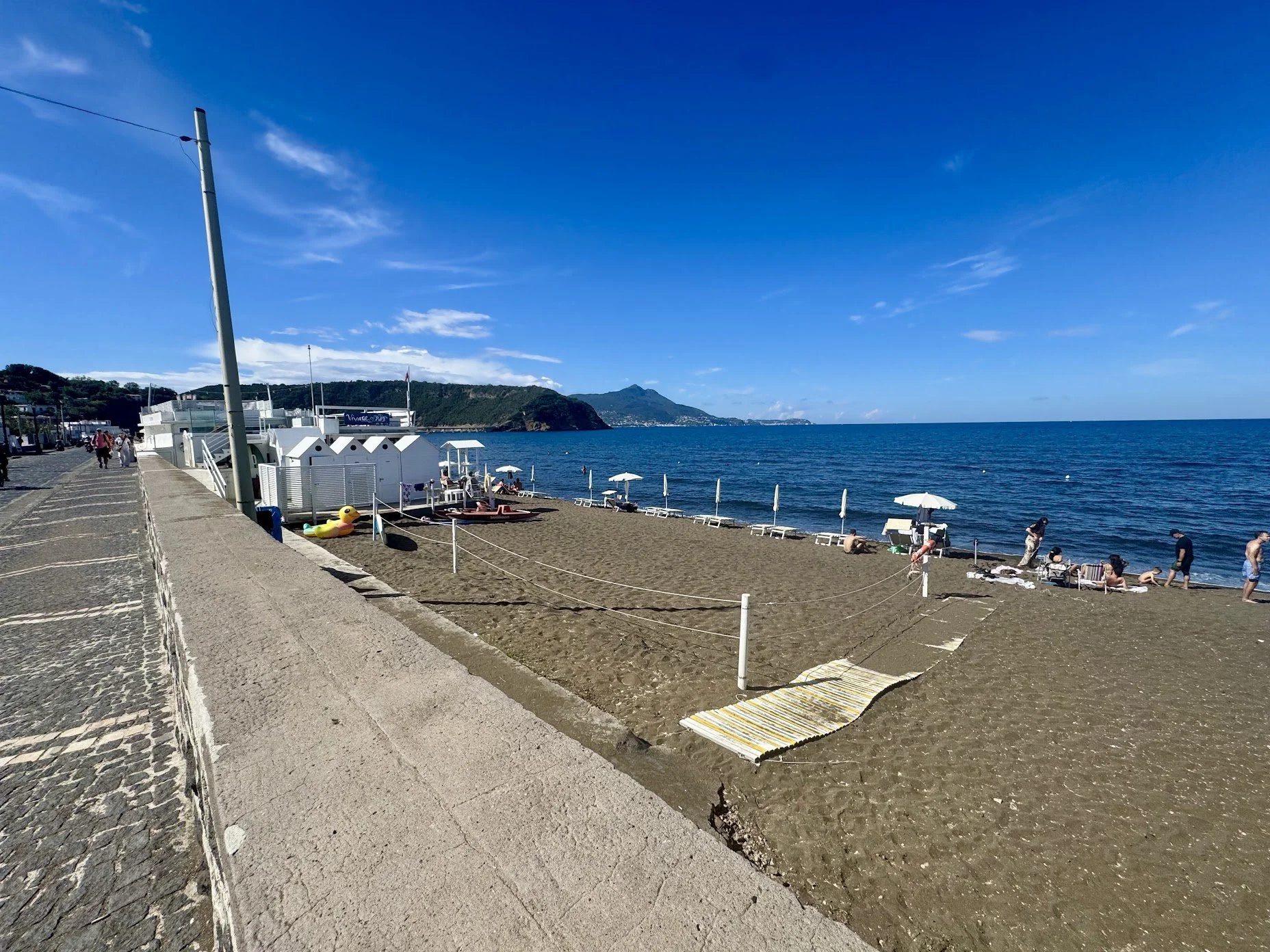 Beach with umbrellas and lounge chairs, shoreline with people, blue sky, and distant hills.