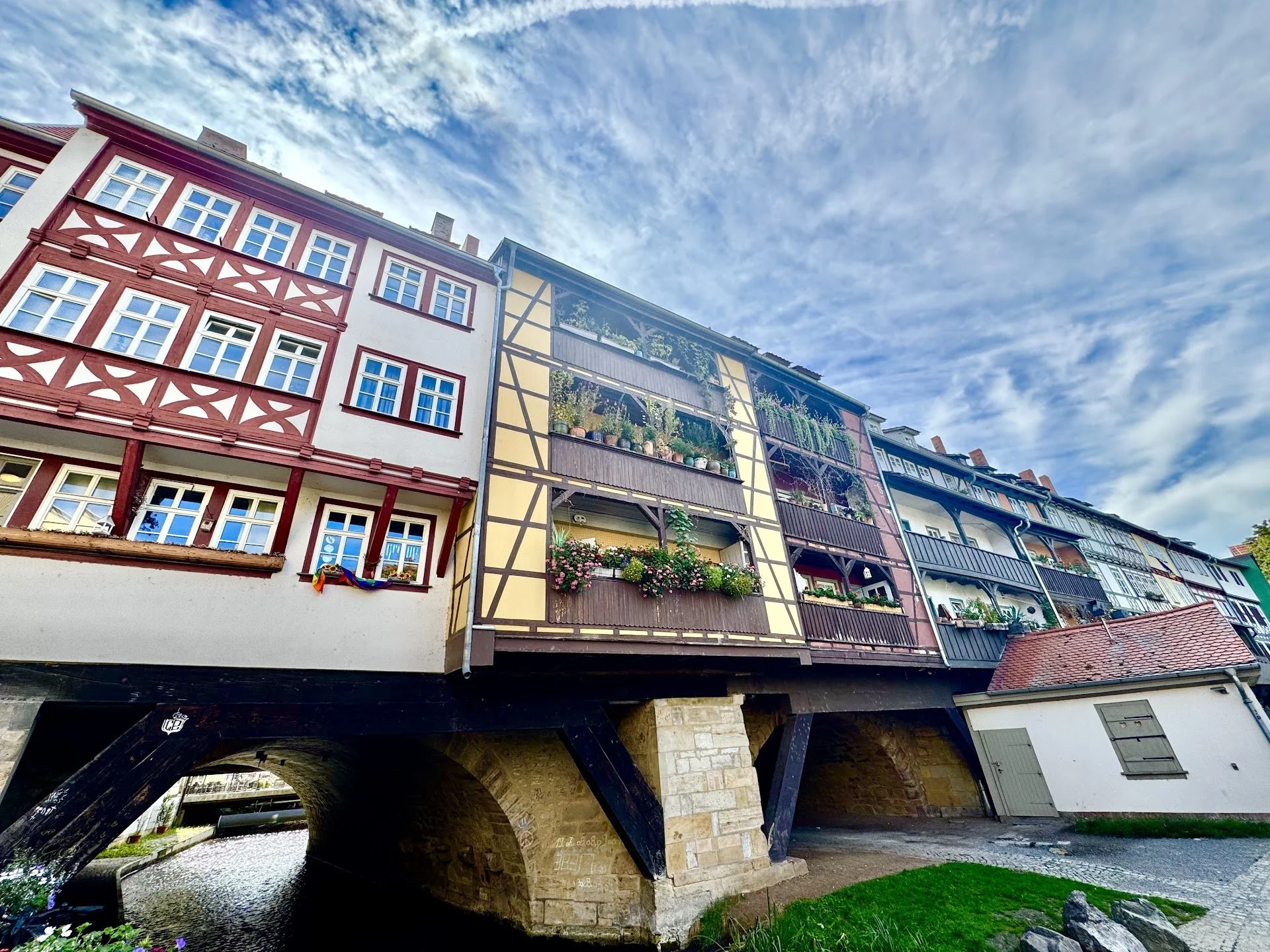Colorful multi-story buildings with decorative facades and balconies, built on a stone bridge arch over a small cobblestone street, under a partly cloudy sky.