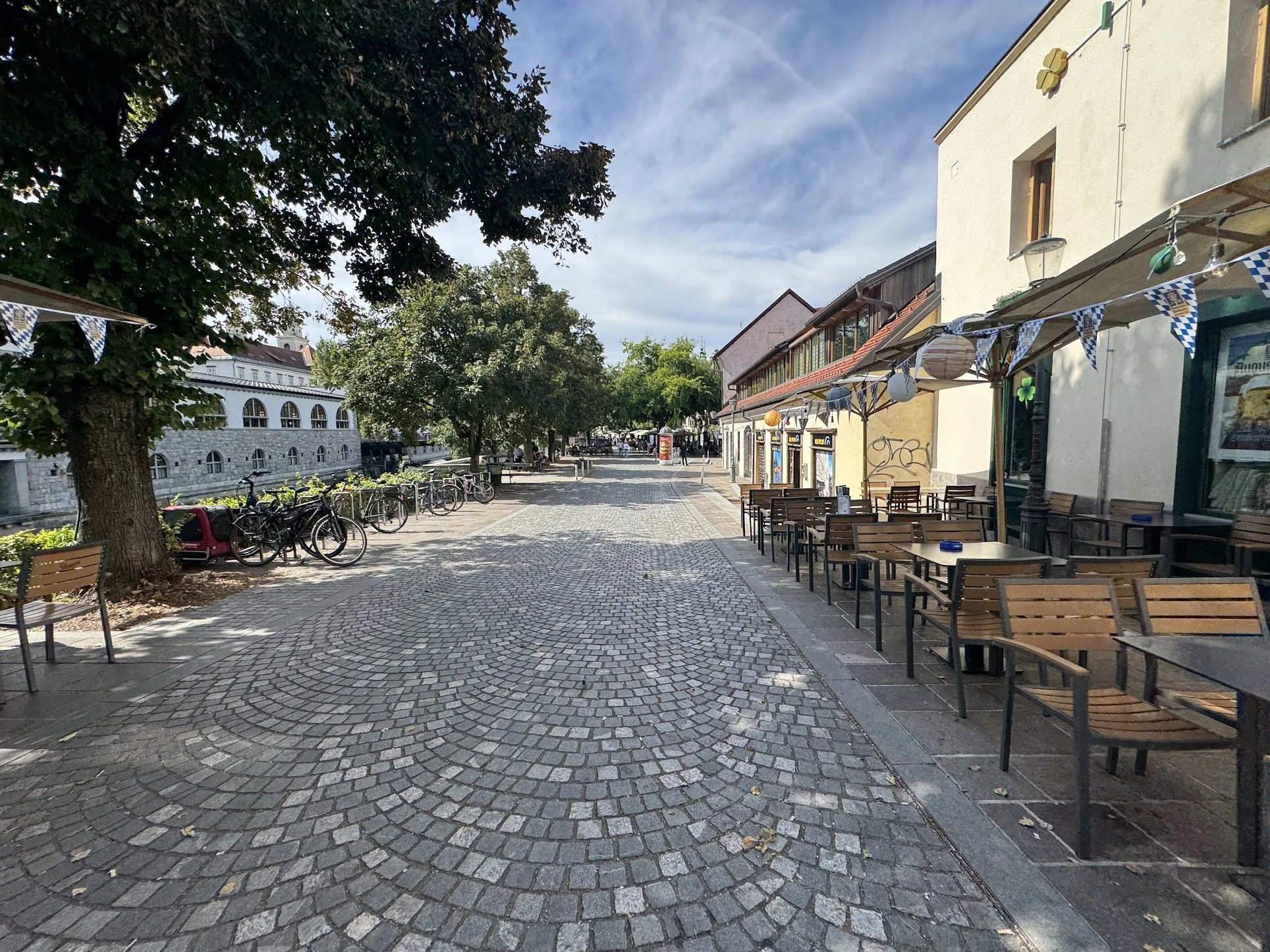 Empty outdoor seating area with chairs and tables, a cobblestone sidewalk, trees lining the street, buildings with overhangs and decorative banners, and a partly cloudy sky.