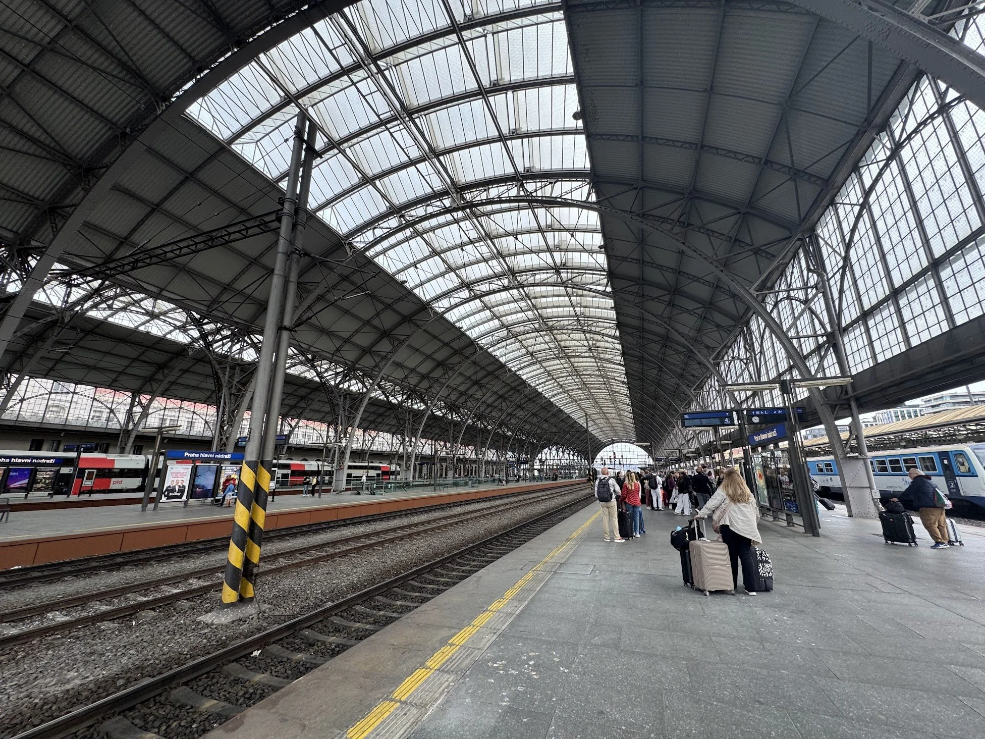 Interior of a large, modern train station with a high arched glass ceiling, train tracks, and platform. Passengers with luggage wait or walk along the platform.