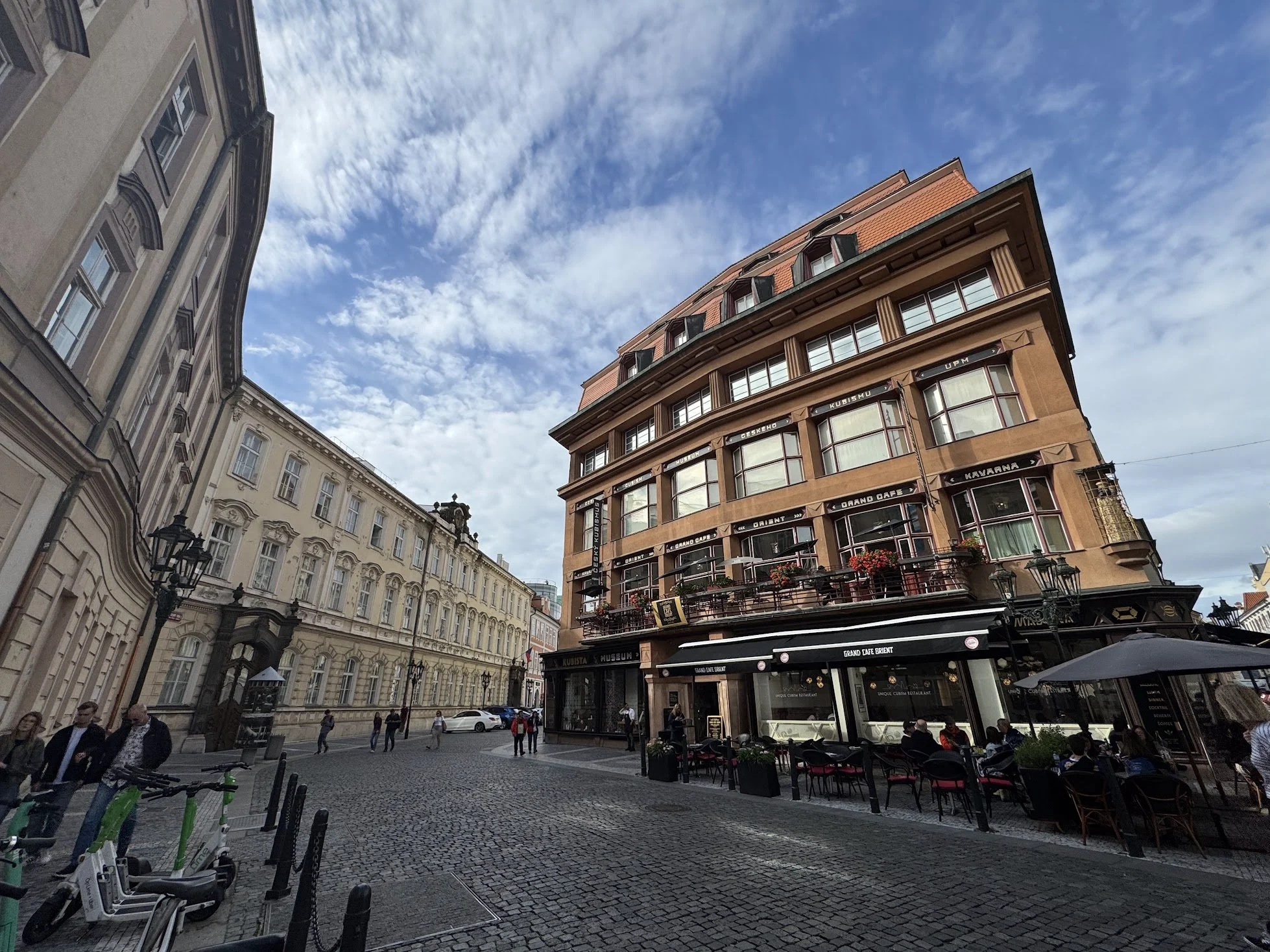 Street scene in a European city with historic buildings, outdoor cafe seating, pedestrians, and cobblestone pavement on a partly cloudy day.