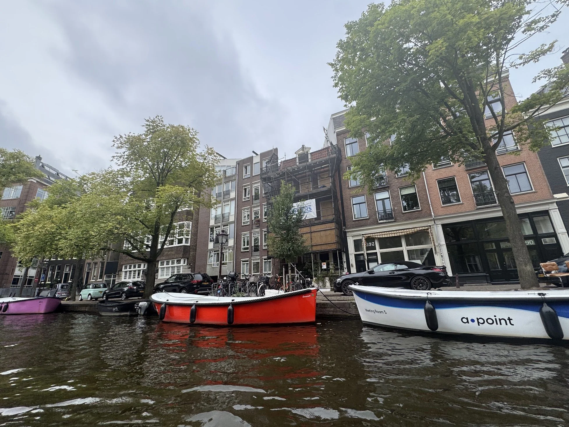 Urban canal scene with boats docked along the water, trees lining the street, and tall apartment buildings with fire escapes against a cloudy sky.