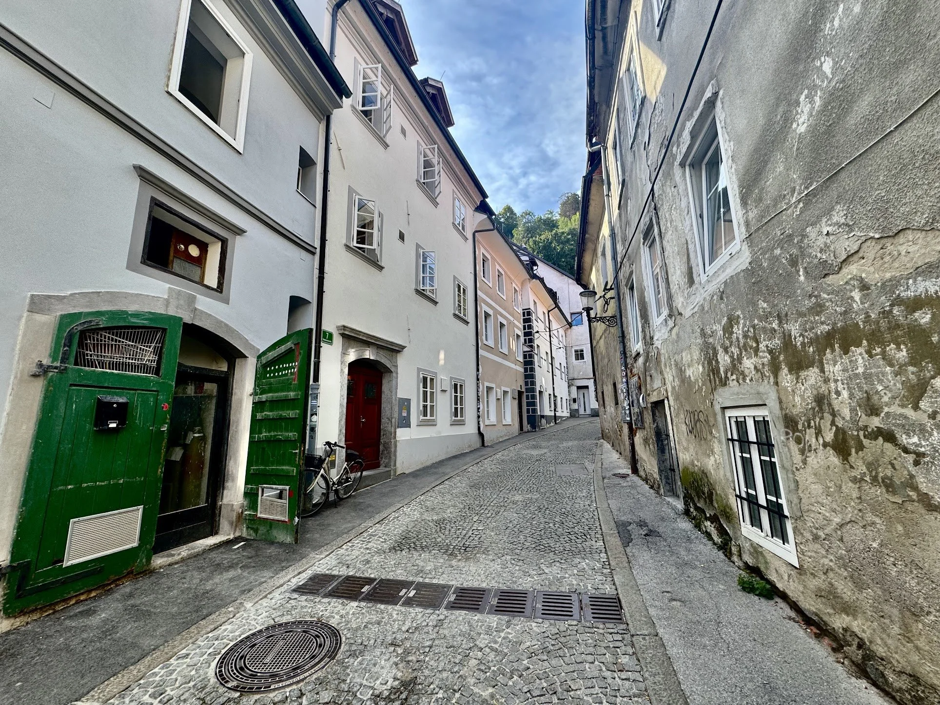 Narrow cobblestone street lined with old white and weathered beige buildings, some with open windows and a green gate on the left side, under a blue sky.