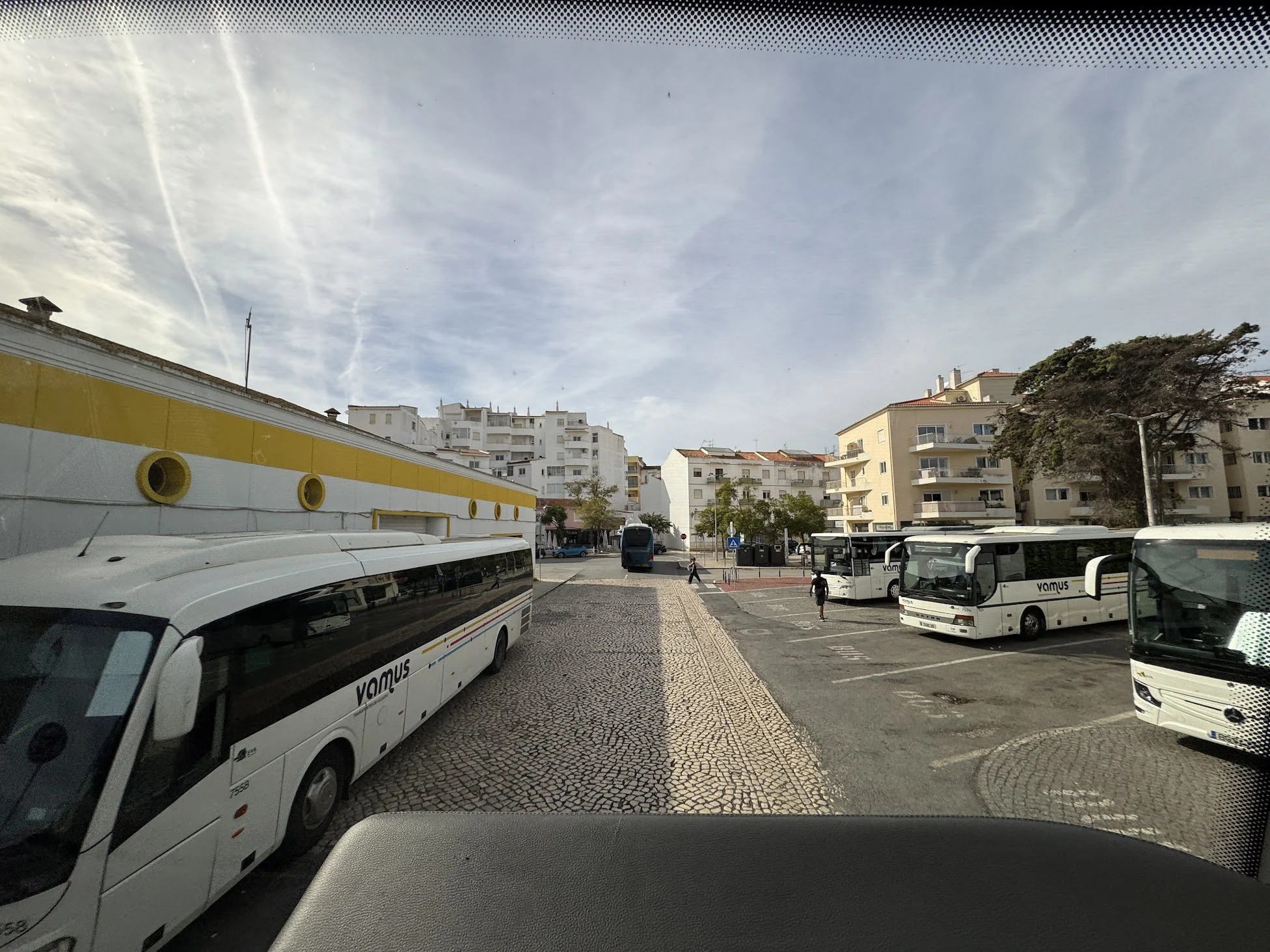 City bus station with multiple white buses parked and people walking around, surrounded by white residential buildings and trees, with a partly cloudy sky overhead.