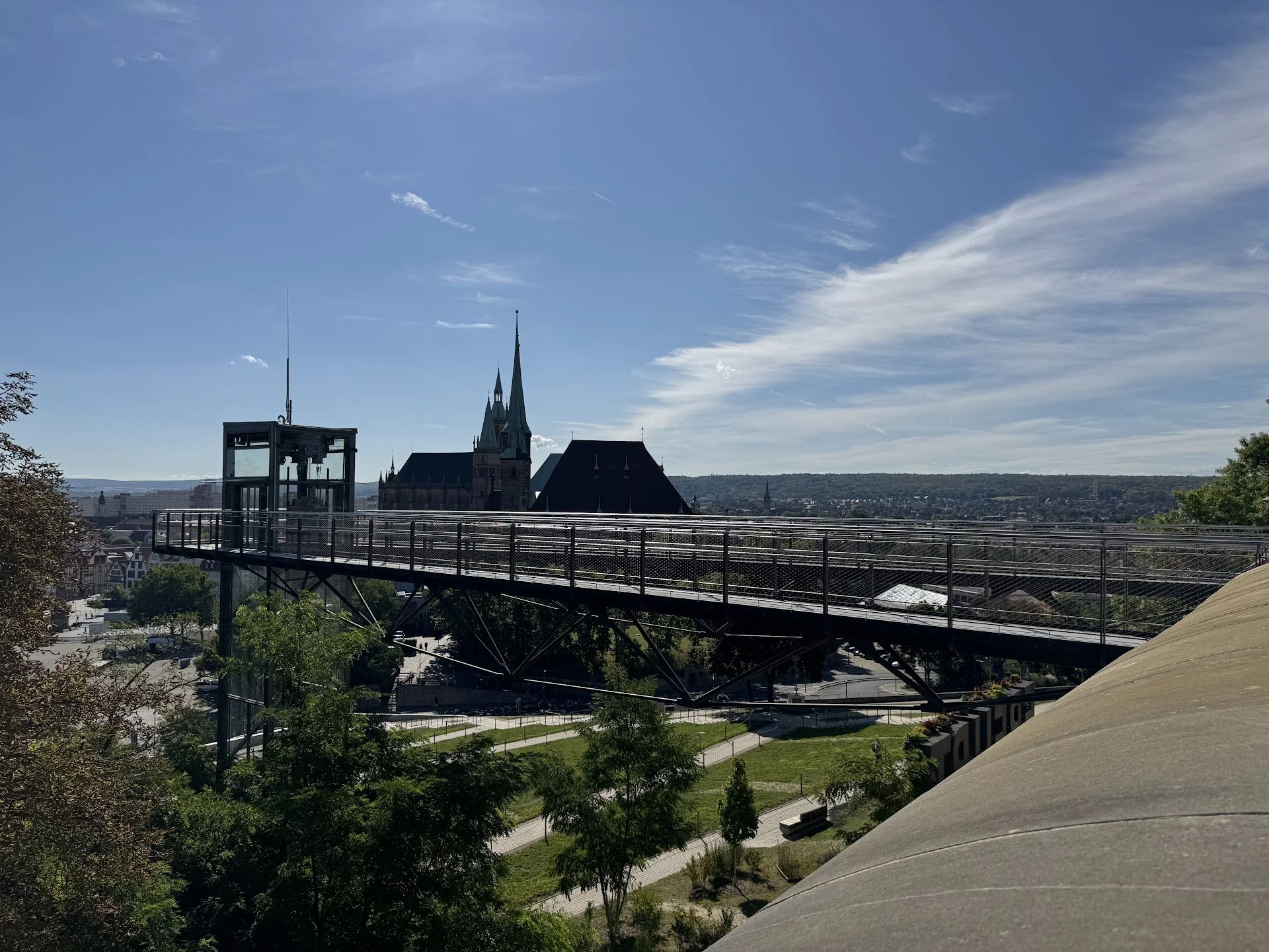 Cityscape view with church steeples, a glass elevator structure, and a railway bridge, under a partly cloudy sky.
