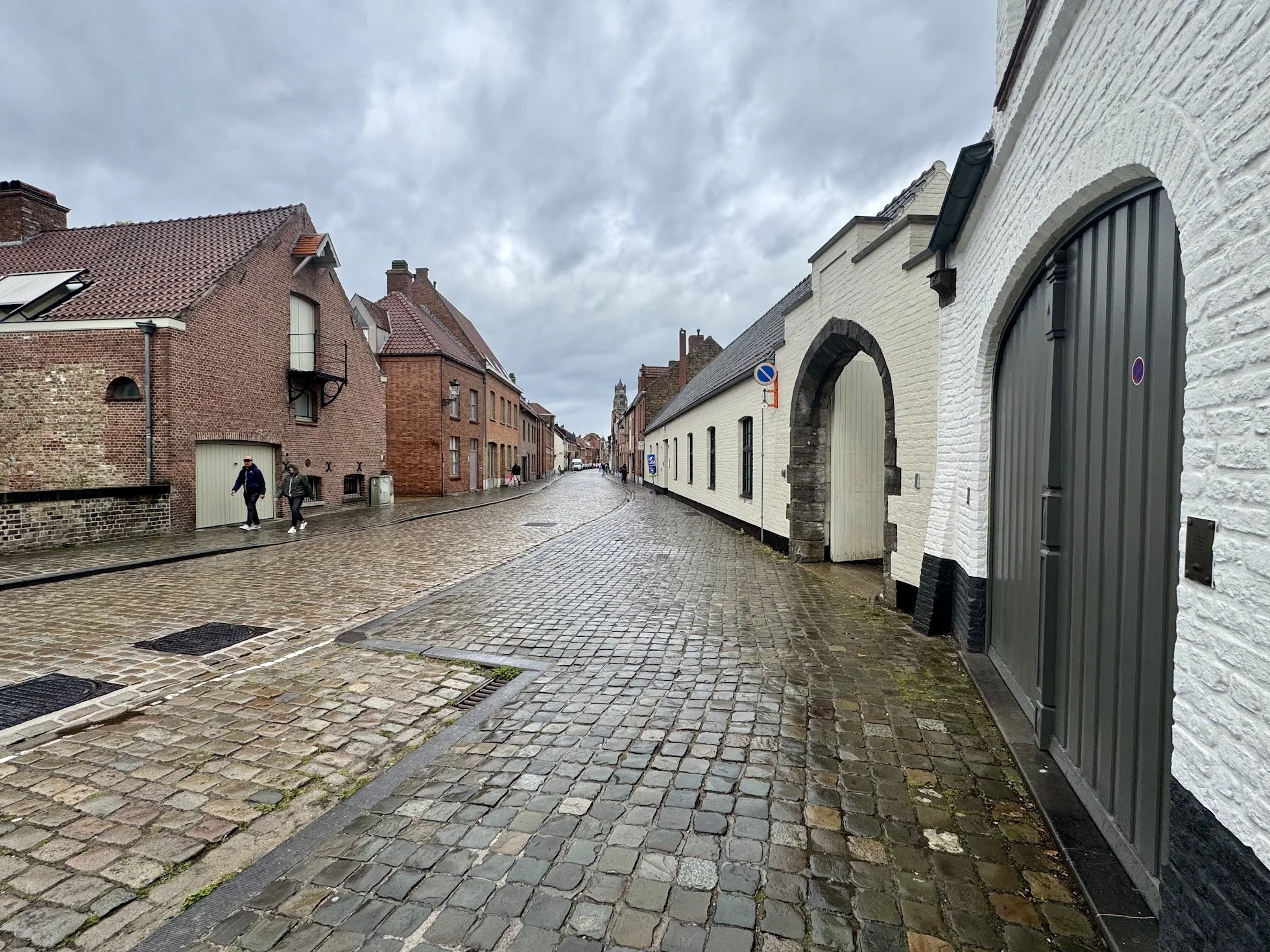 A cobblestone street in a European city with buildings on either side, some made of red brick and others painted white, under a cloudy sky.