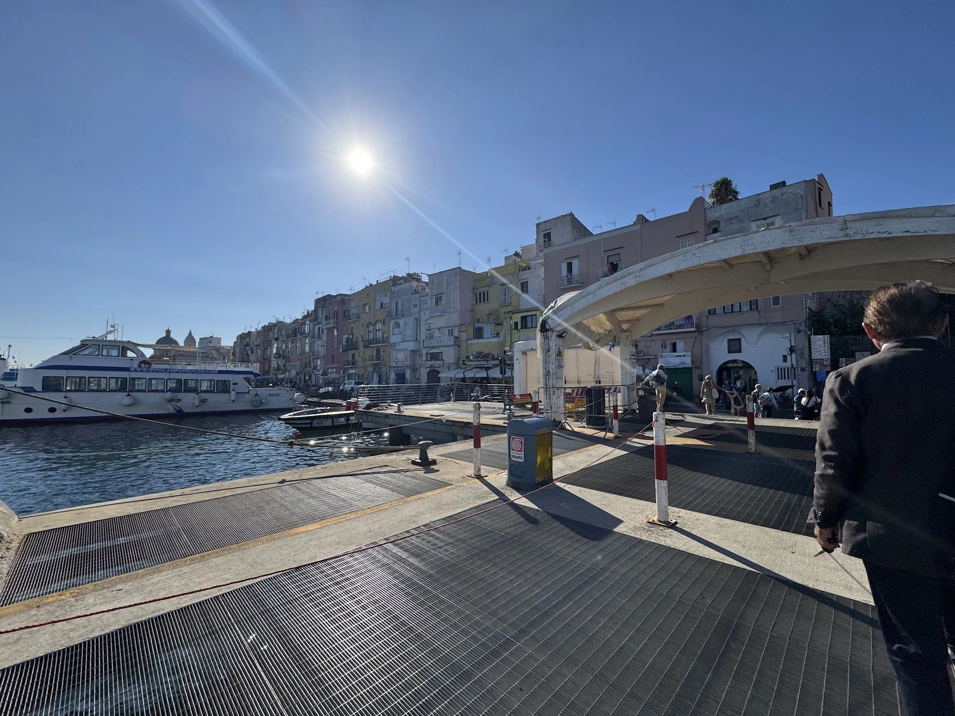 Bright sunny day at a harbor with colorful buildings lining the waterfront. A large yacht is docked in the water, and people are walking along the pier. A man in a suit is in the foreground, and there are boats and a bridge structure over the water.