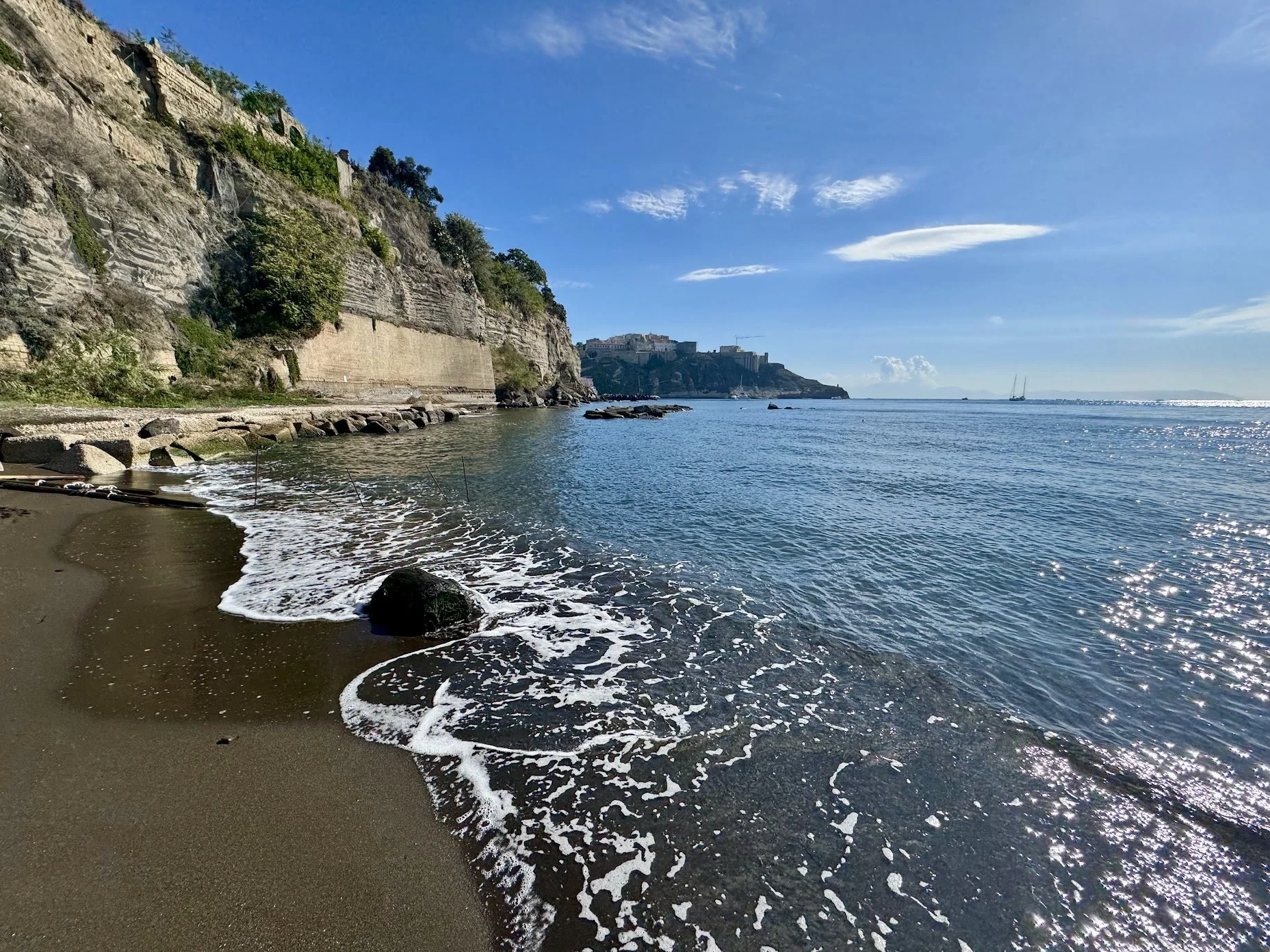A beach with dark sand and small rocks, waves gently crashing onto the shore, steep cliffs with greenery and ruins, and a distant castle on a hill, under a blue sky with scattered clouds.