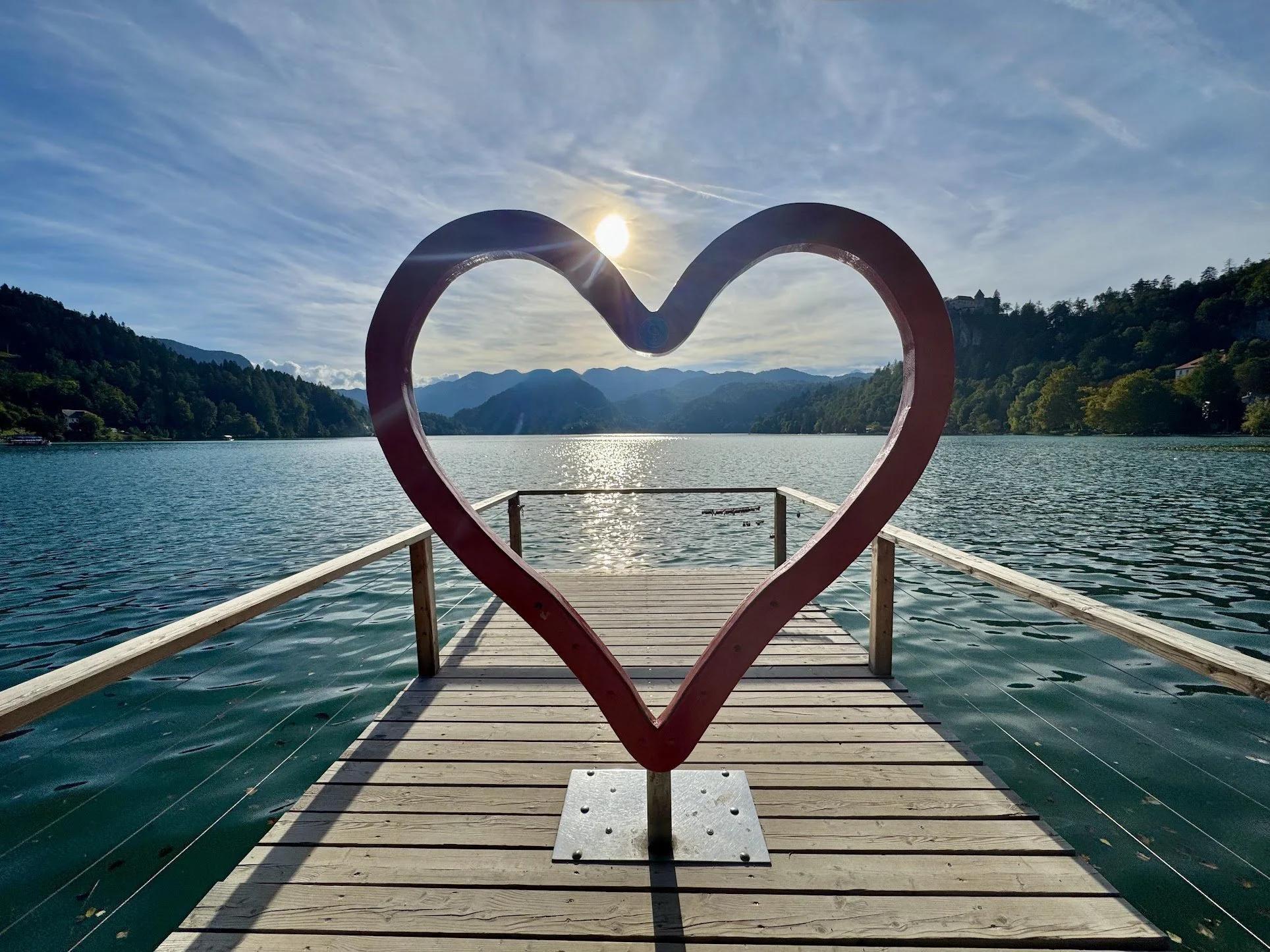A heart-shaped metal sculpture on a wooden dock overlooking a lake with forested mountains in the background and the sun setting or rising behind the sculpture.