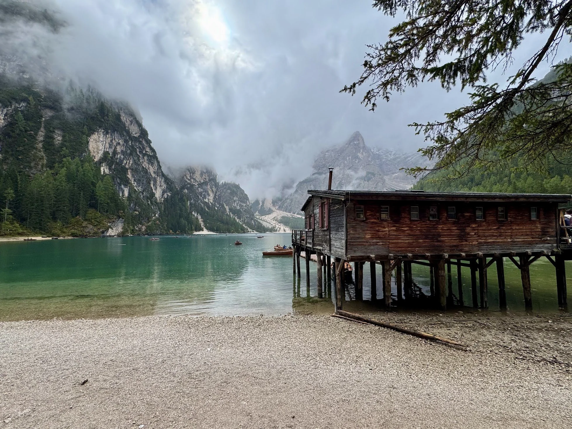 A wooden cabin on stilts by a mountain lake with kayakers, surrounded by tall pine trees and cloudy sky.