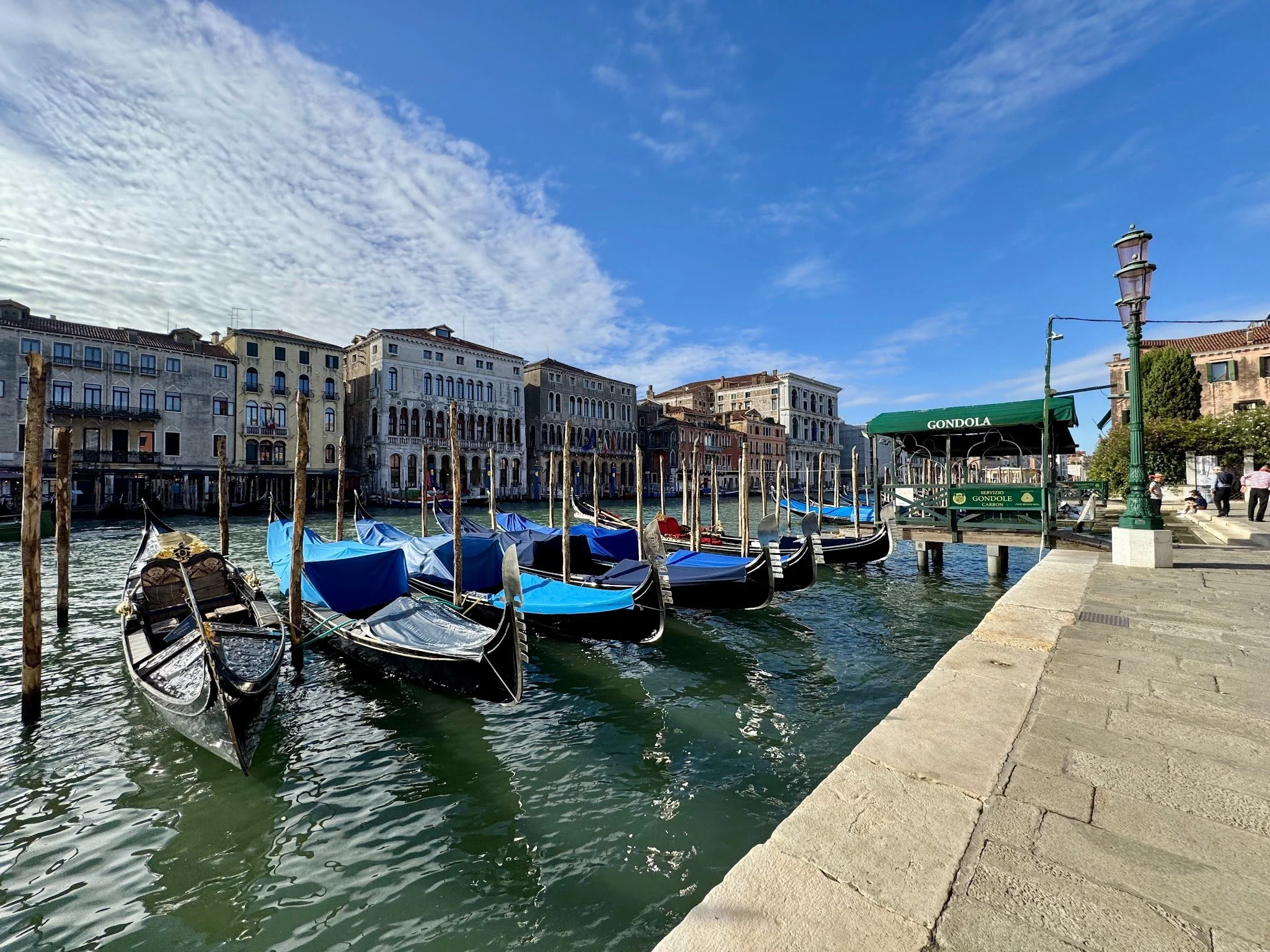 Gondolas docked along a canal in Venice, Italy, with historic buildings in the background and a partly cloudy blue sky overhead.