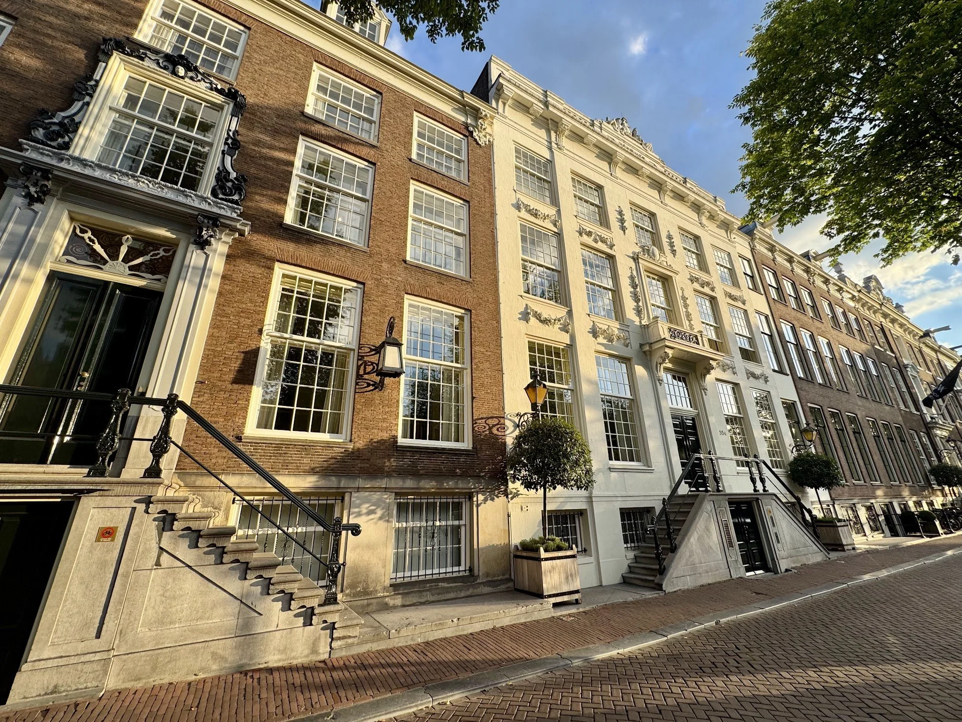 Street view of multi-story brick and white buildings with large windows, front stairs, and decorative architectural details, under a partly cloudy sky with trees.