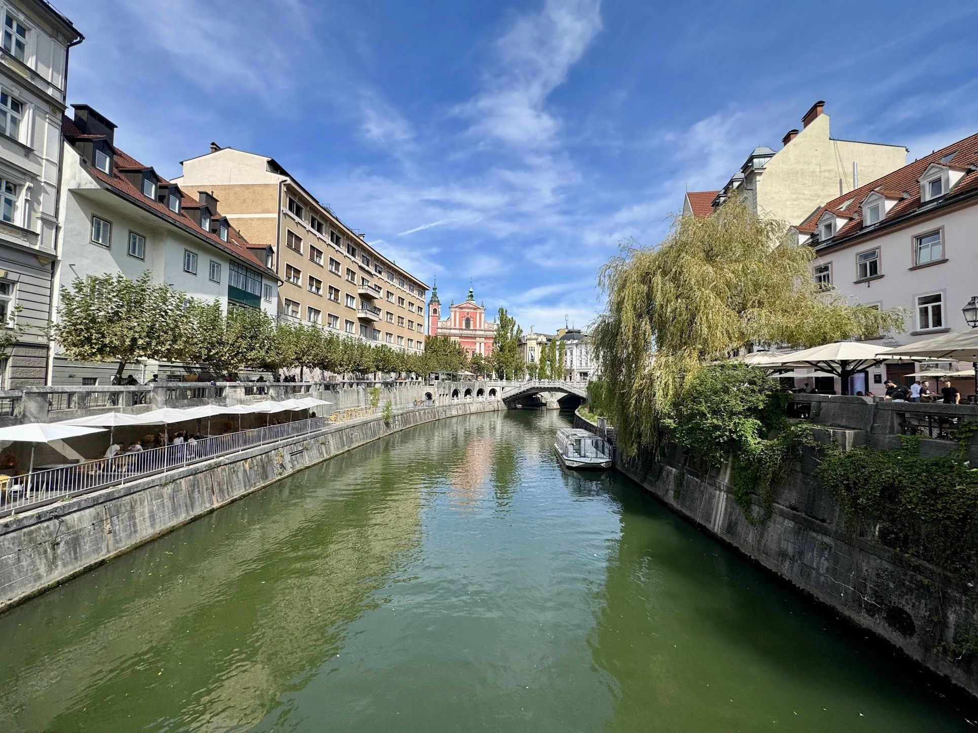 A canal lined with European-style buildings on both sides, featuring trees, outdoor cafes, and a bridge with a pink church in the background under a blue sky with scattered clouds.
