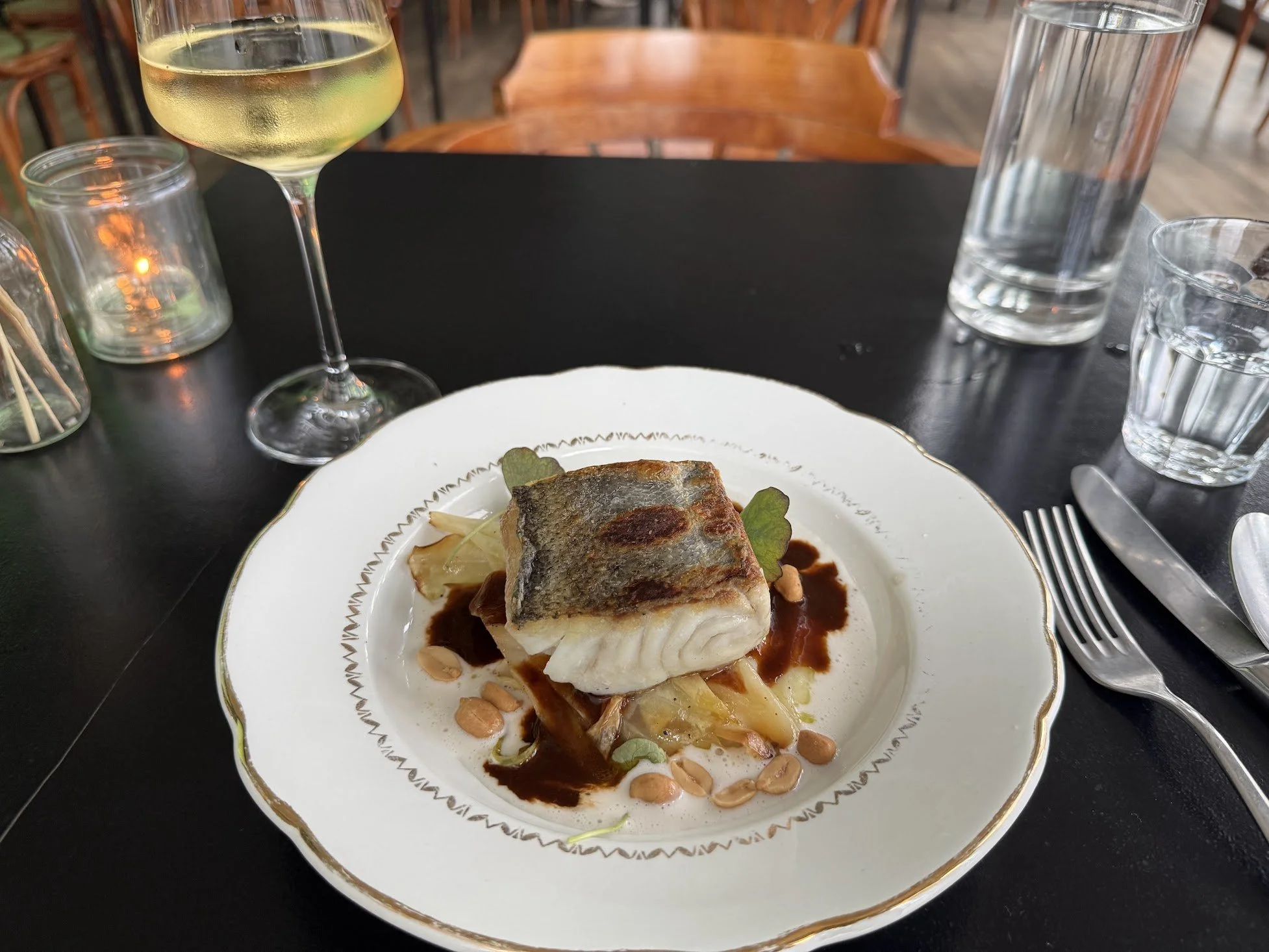 A plated serving of cooked fish with a browned crust, topped with sauce, served over vegetables and garnished with microgreens, on a white decorative plate. The table also holds a glass of white wine, some glasses of water, and a candlestick, in a re