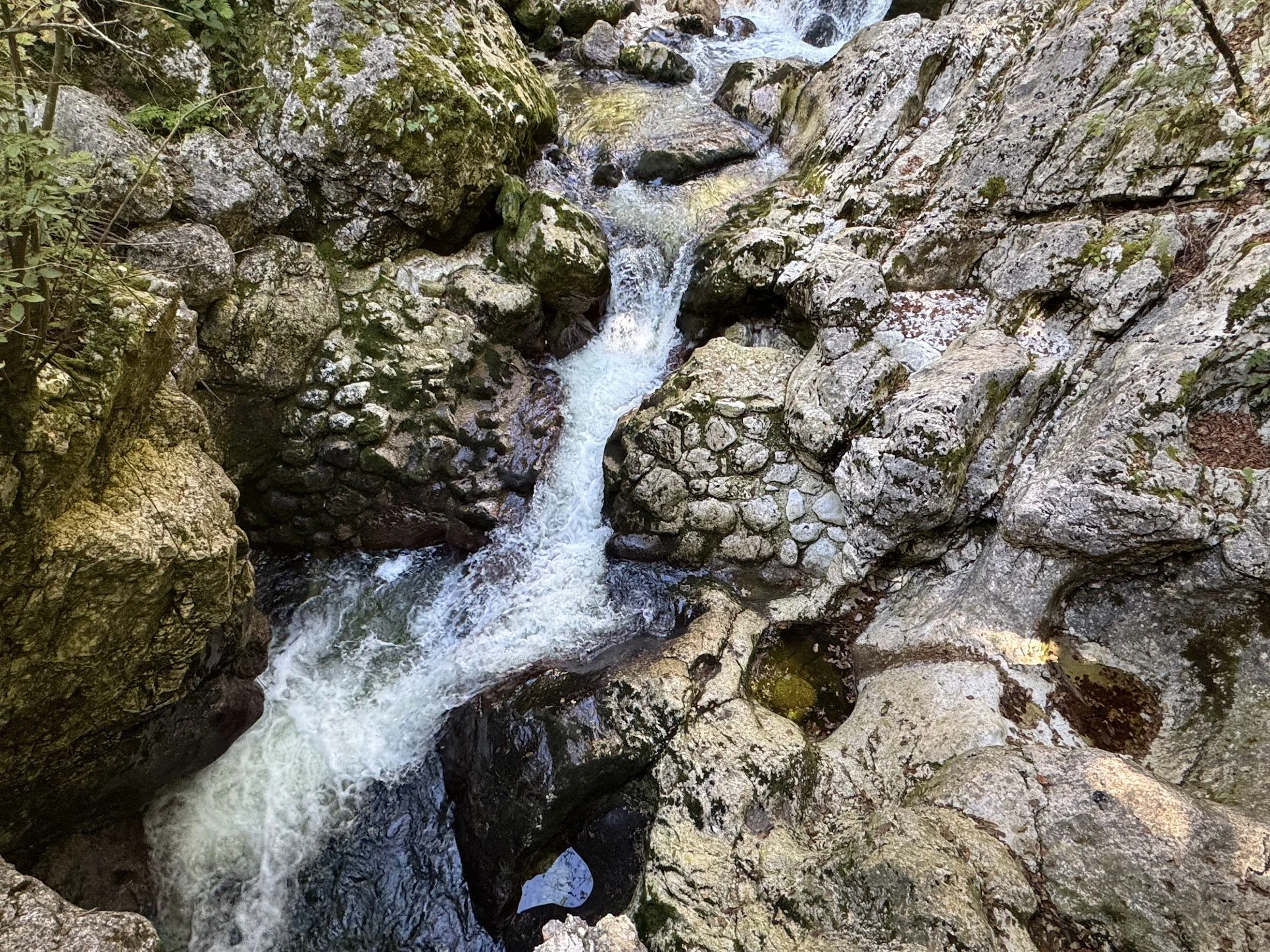 A small mountain stream flowing between large moss-covered rocks and boulders in a narrow rocky canyon surrounded by greenery.