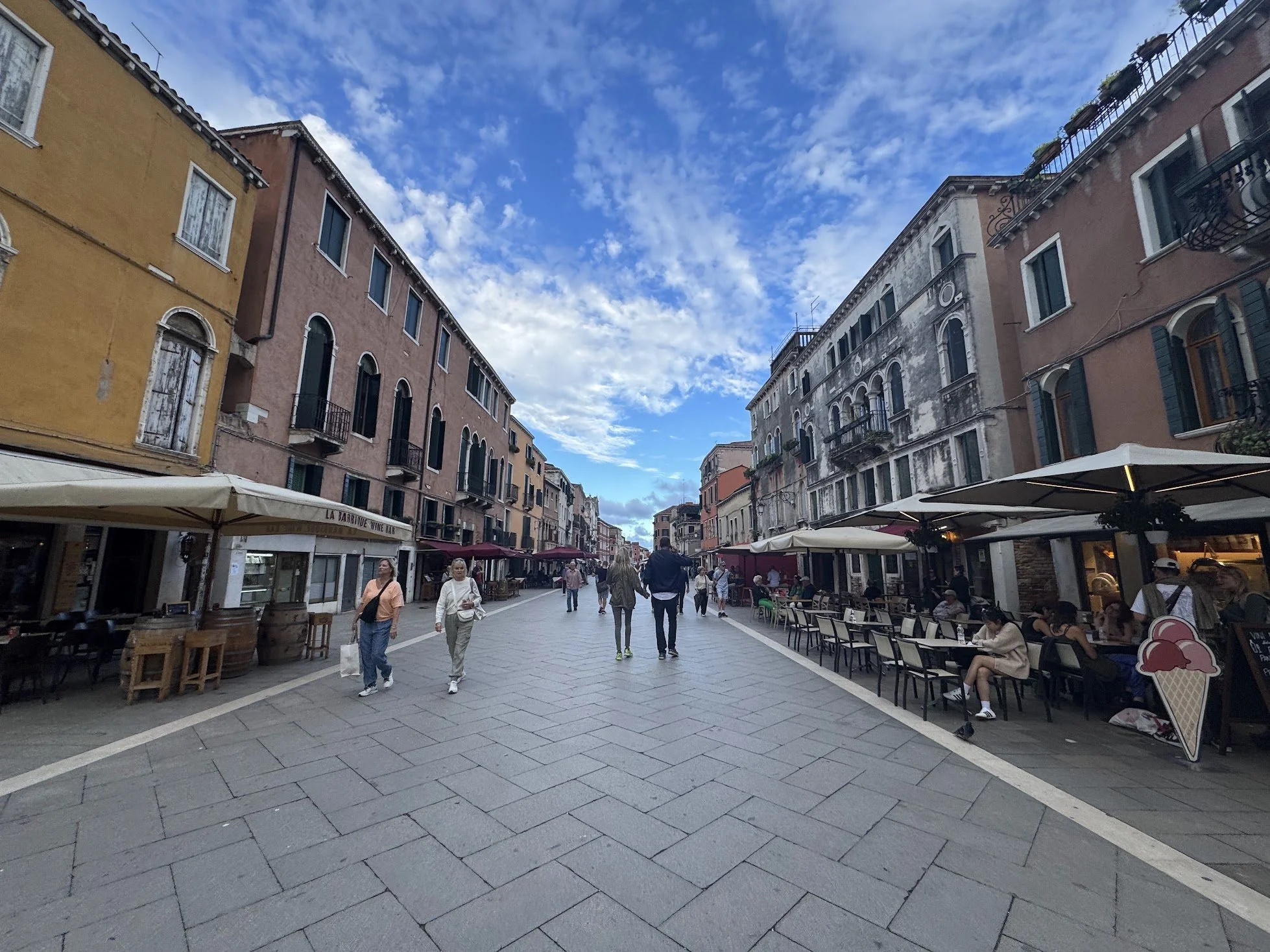 A bustling outdoor market street with colorful buildings, outdoor seating, and people walking and sitting at cafes, under a partly cloudy sky.