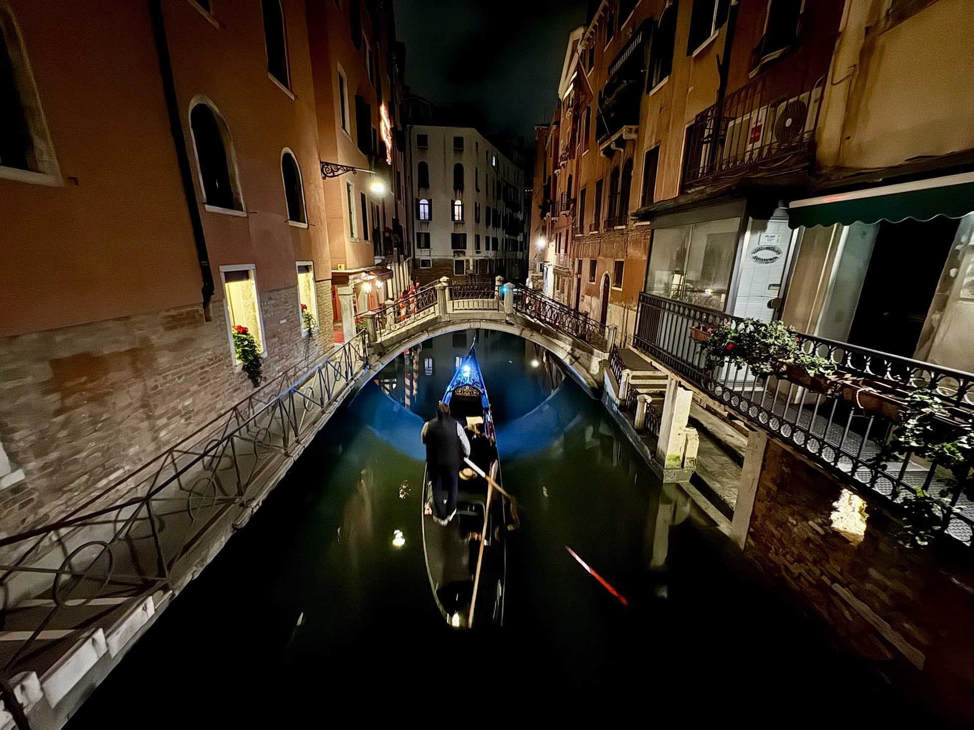 Nighttime scene of a canal in Venice with a gondola being steered by a gondolier, surrounded by colorful buildings with lit windows and balconies.
