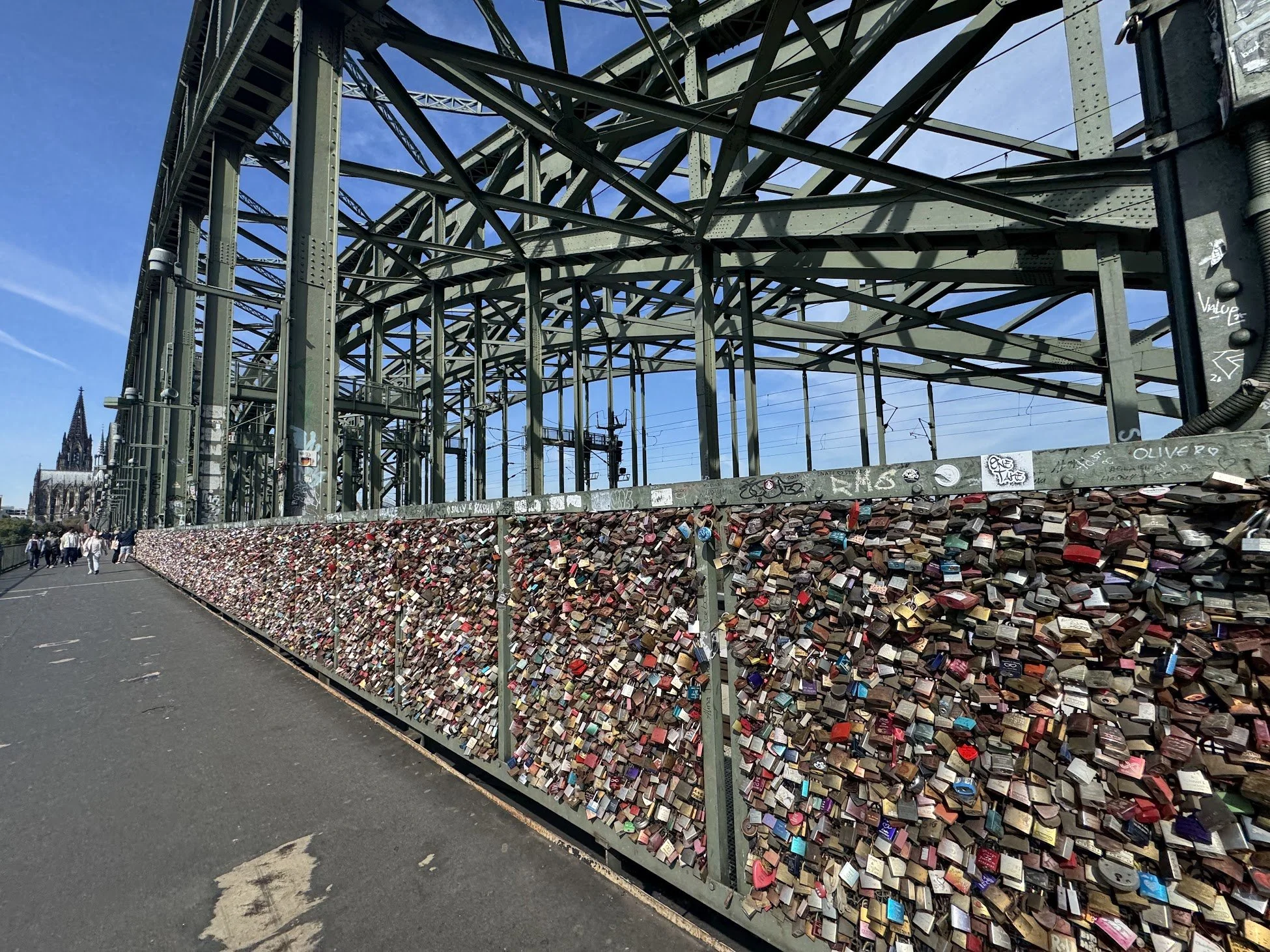 A bridge with numerous padlocks attached to its railing, with a large cathedral in the background and a group of pedestrians walking on the paved area beneath.
