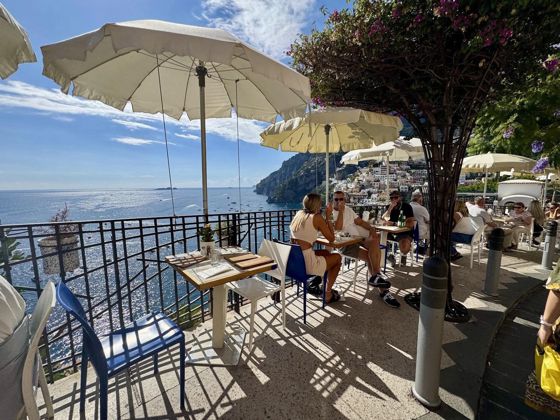 A seaside restaurant terrace with people dining under large beige umbrellas, overlooking the ocean and a cliffside town, with clear blue sky and sunlight reflecting on the water.