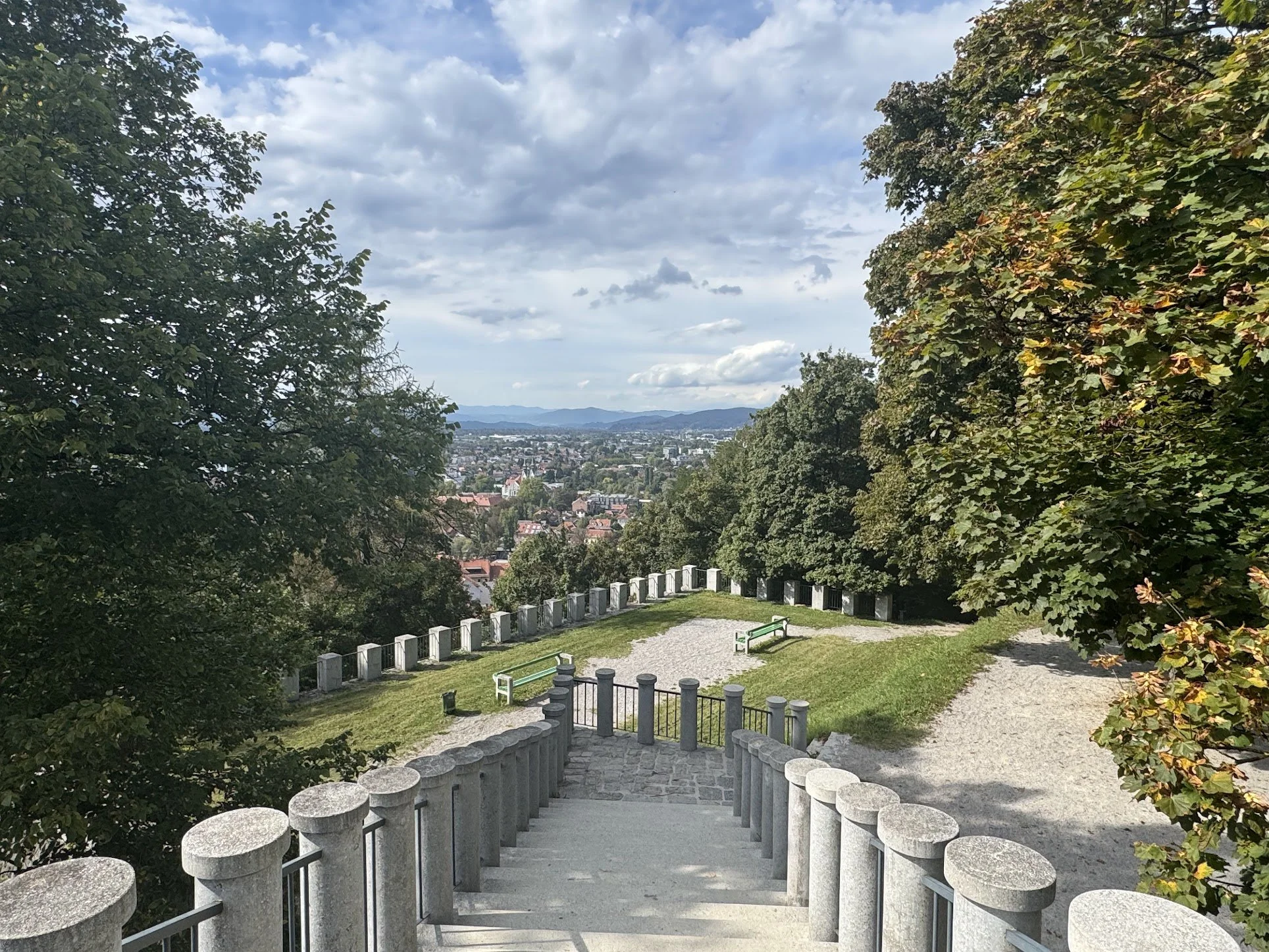 A scenic view from a hilltop park with stone steps leading down, surrounded by trees with green leaves, overlooking a cityscape with houses, buildings, and mountains in the distance under a partly cloudy sky.