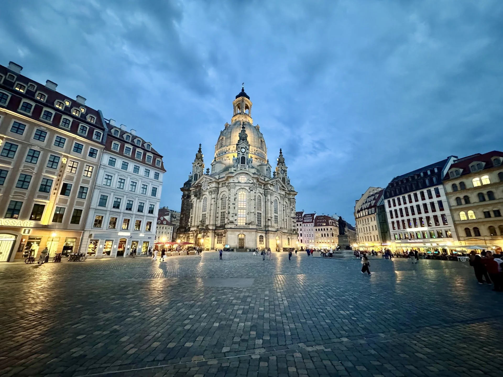City square at dusk with a historic church surrounded by modern buildings and people walking.