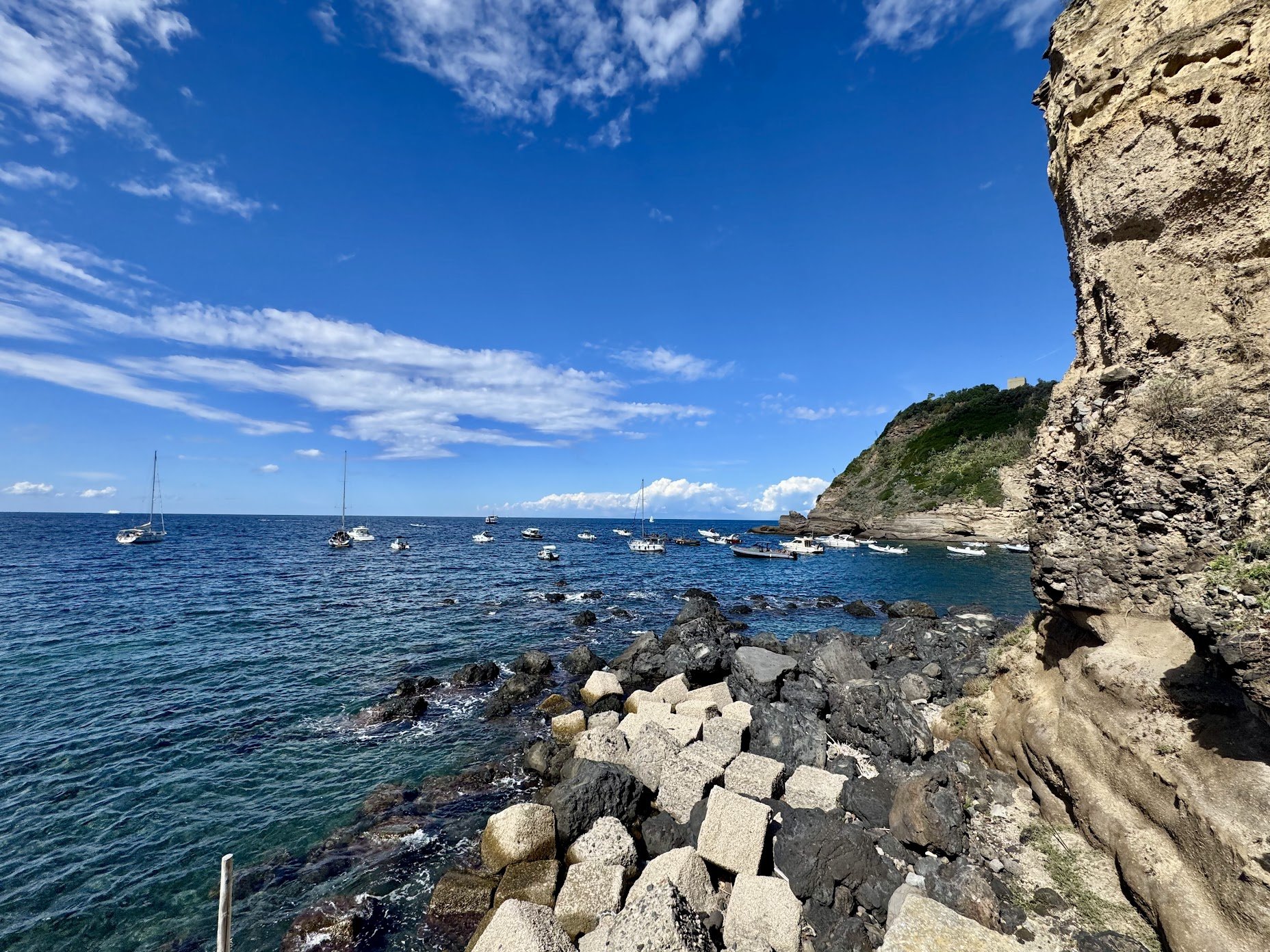 A scenic ocean view with a rocky shoreline in the foreground, sailboats and motorboats on the water, a large rocky cliff on the right, and a partly cloudy sky above.