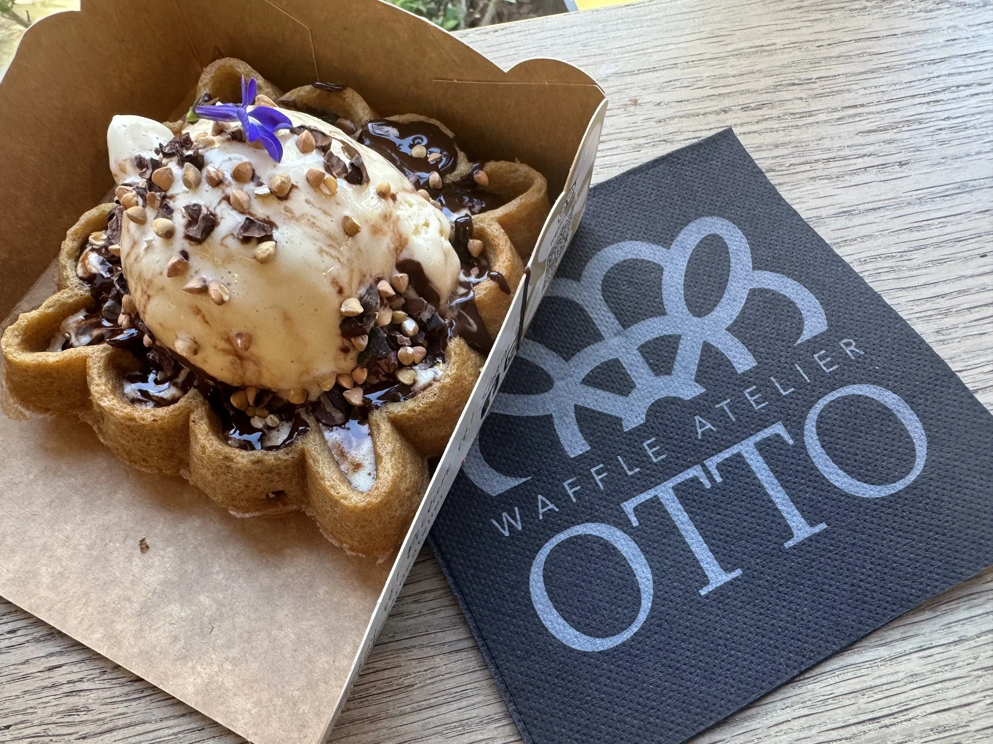 A Belgian waffle topped with vanilla ice cream, chocolate syrup, chopped nuts, and a violet edible flower, placed on a piece of parchment paper on a wooden table, alongside a black napkin with a geometric pattern.