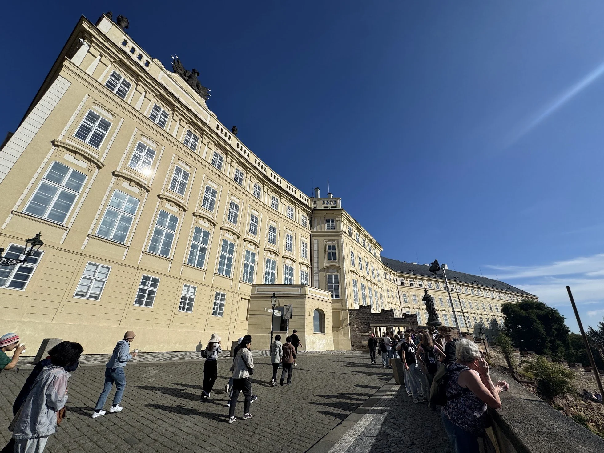 A large yellow historic building with numerous windows and statues on the roof. People are walking and standing outside in a sunny area.