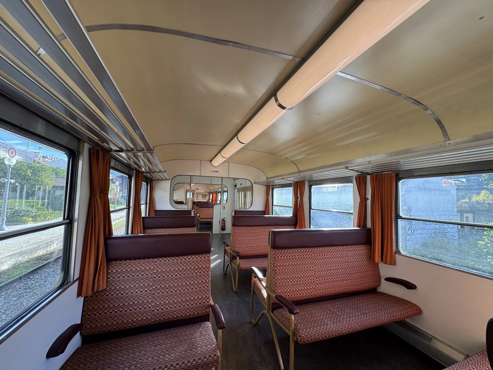 Interior of a vintage train car with red checkered seats, large windows with orange curtains, and a wooden-paneled ceiling with a central light fixture.