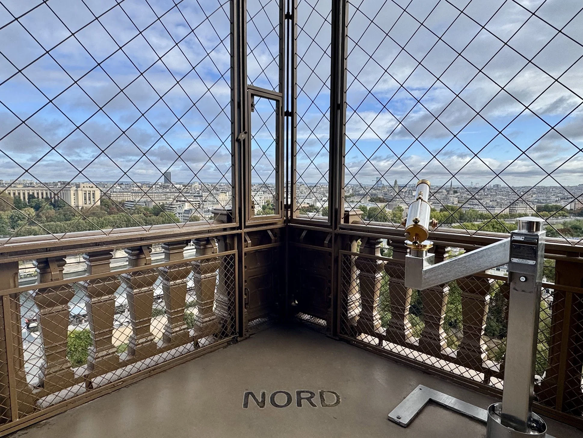 View from an observation deck with a metal railing and a telescope, overlooking a cityscape with various buildings and greenery under a cloudy sky.