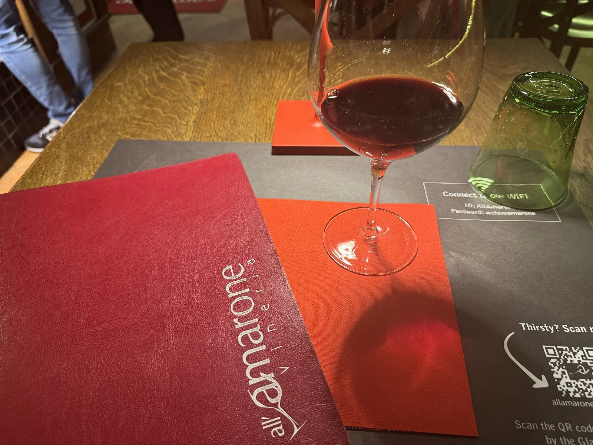 A partially filled glass of red wine on a wooden table with red and gray paper mats underneath. A green glass tumbler is nearby, and a red menu is partially visible in the foreground.