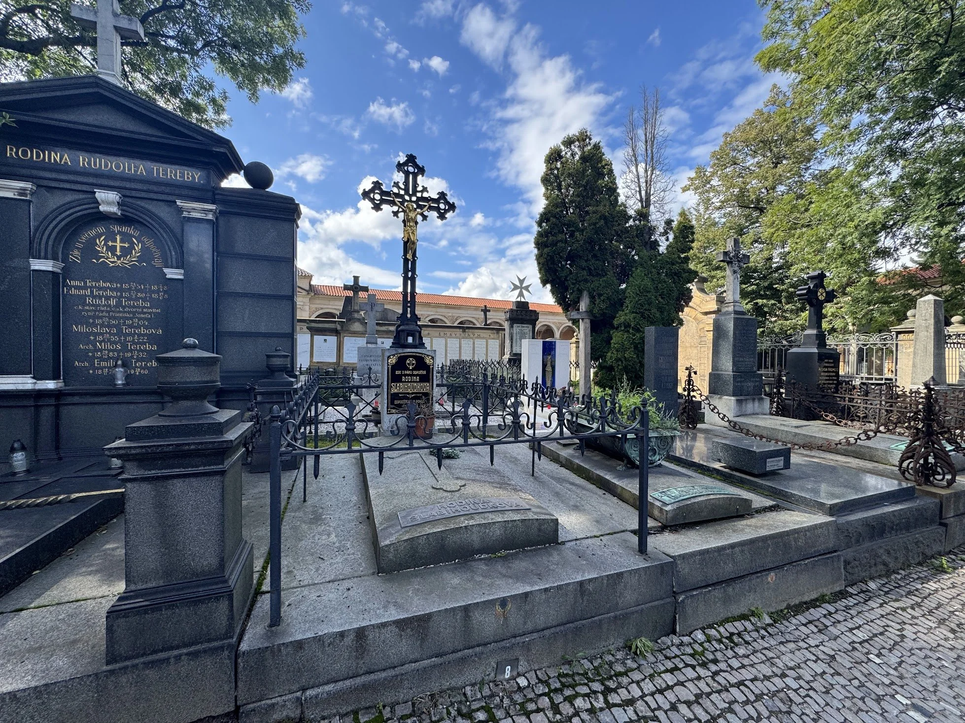 A cemetery with various tombstones and crosses surrounded by trees under a partly cloudy sky.