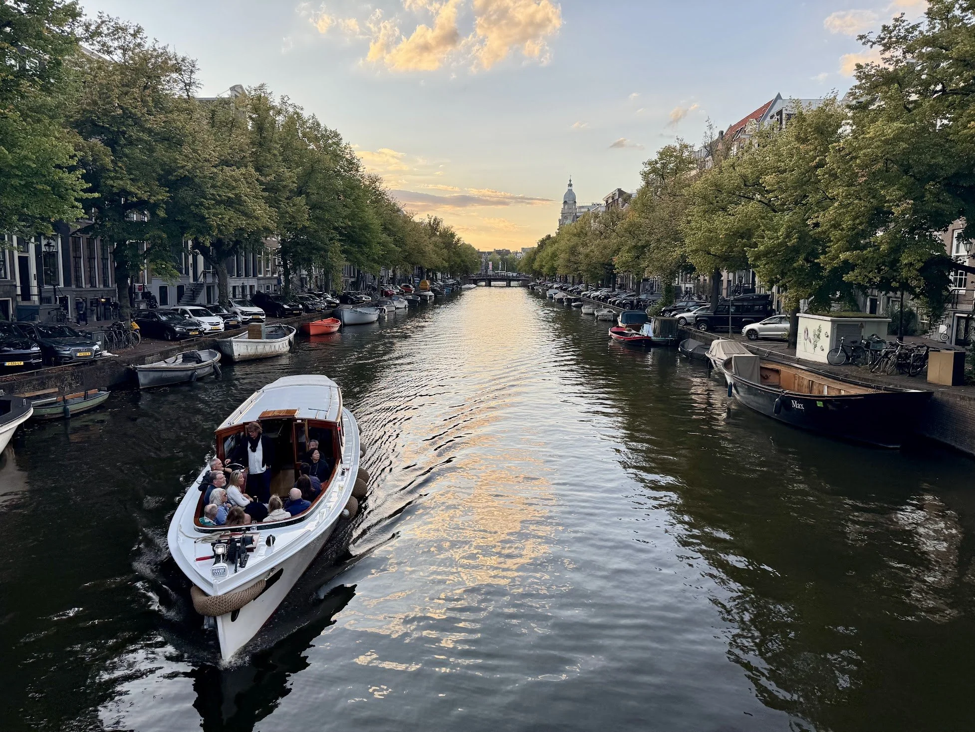 A canal lined with trees and parked cars on both sides, with boats docked along the edges, and a boat carrying tourists in the foreground at sunset.