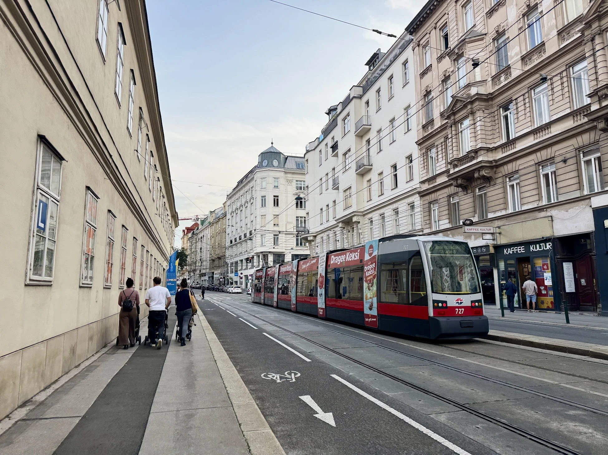 A city street scene with a tram moving along tracks and people walking on the sidewalk, including a woman pushing a stroller. Buildings line both sides of the street, with tram wires overhead and a bike lane marked on the road.