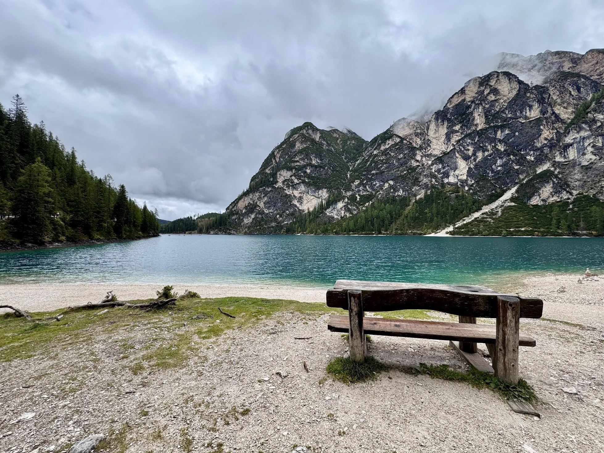Scenic mountain landscape with a lake, forested shoreline, rocky mountains, cloudy sky, and a wooden bench in the foreground.