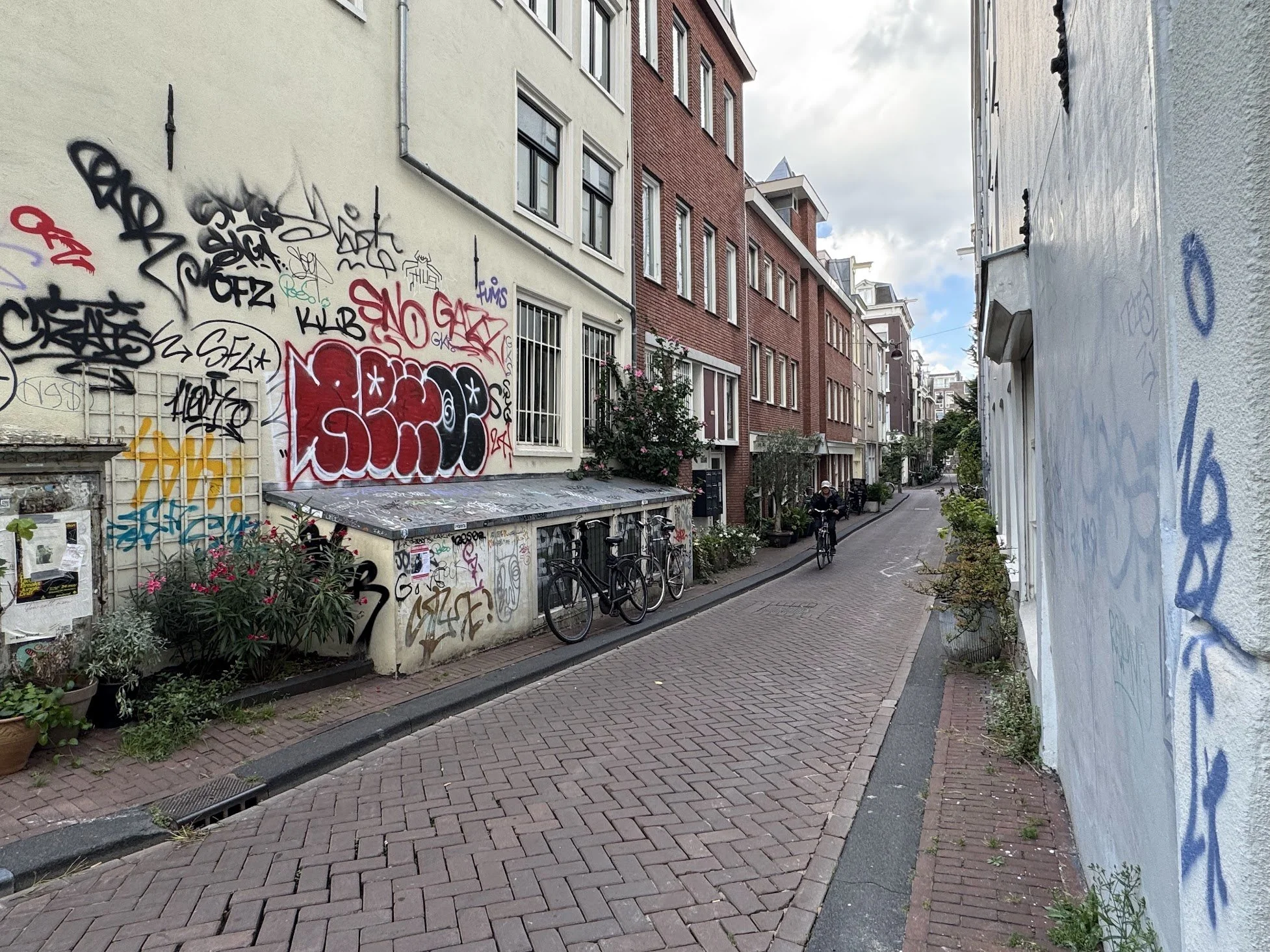 A narrow urban street with brick and stucco buildings on either side, some with graffiti, and bicycles parked near plants. A person is cycling down the street.