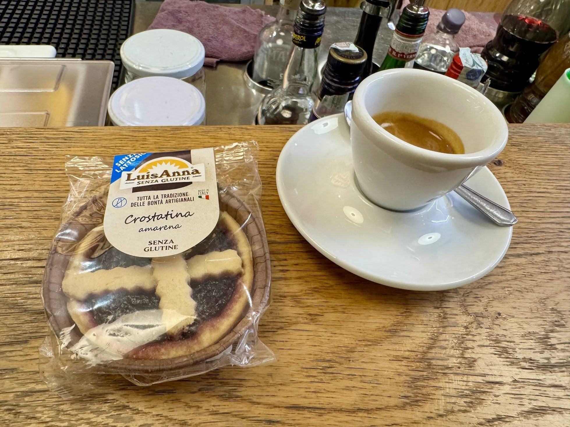 A partially consumed cup of espresso on a white saucer with a metal spoon, a package of gluten-free cookies labeled "Crostatina amarena," and various condiments, salt, and pepper shakers on a wooden table.